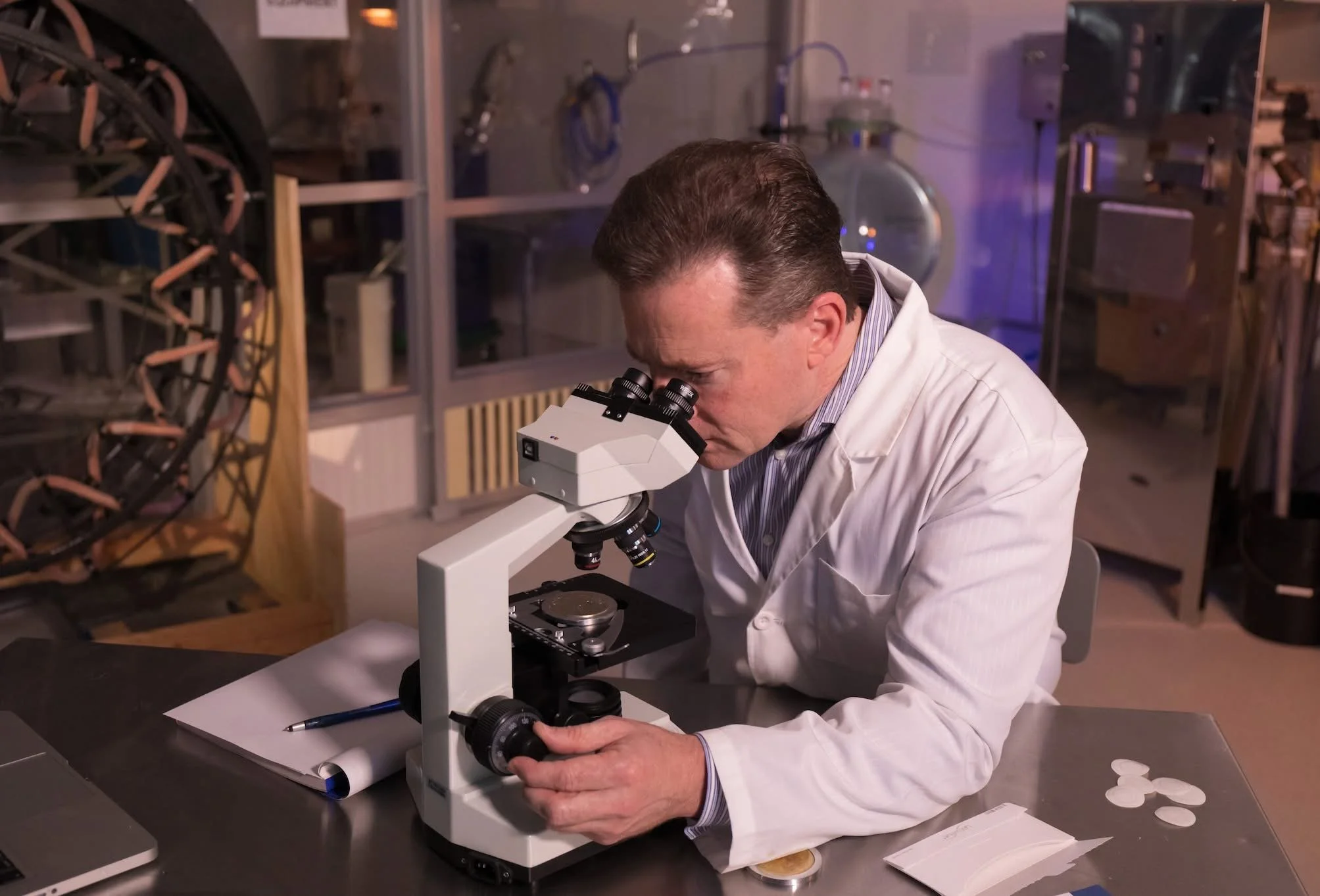 A David Schmidt in a white lab coat examining a slide under a microscope in a laboratory setting.
