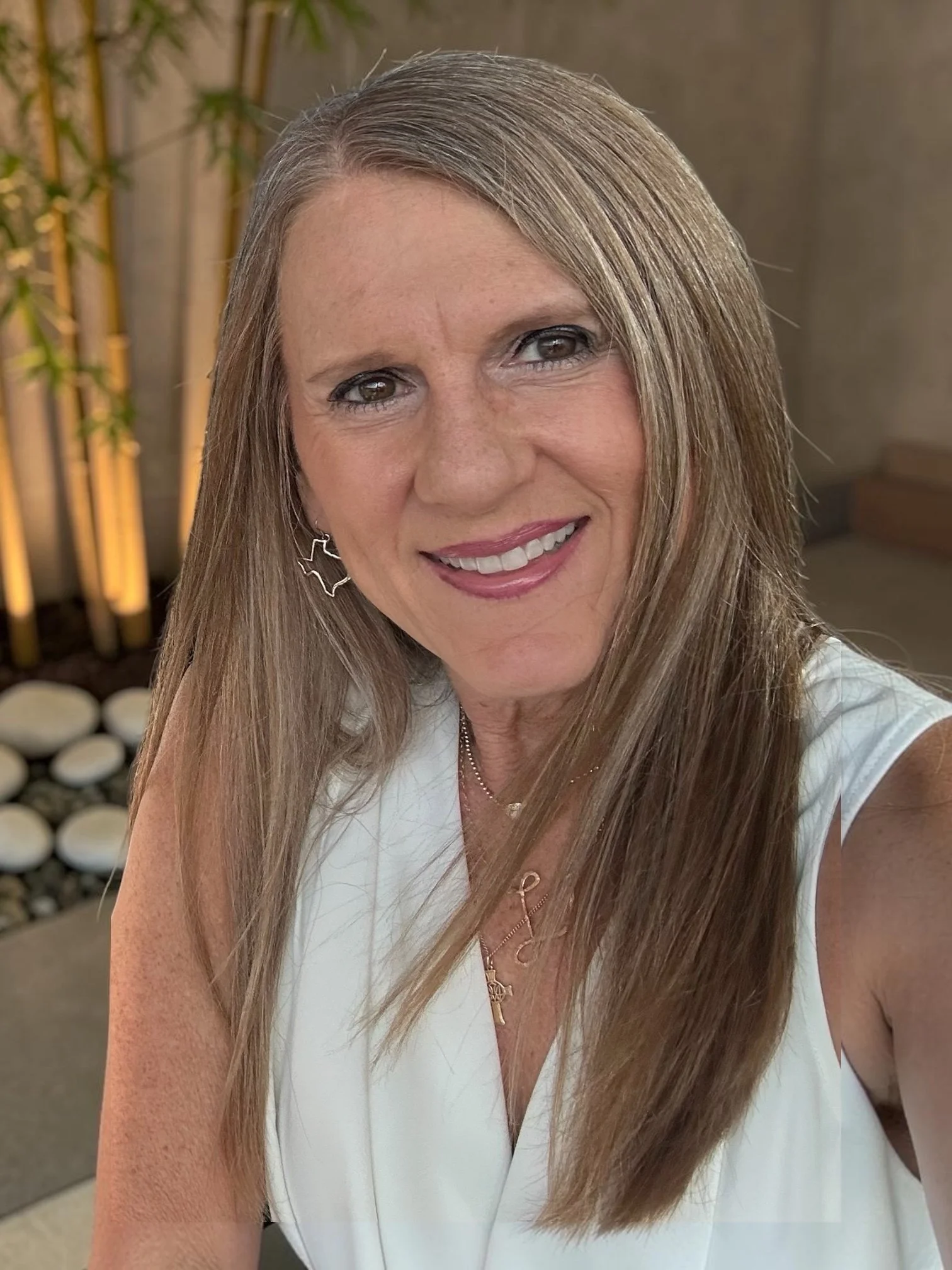 Leah with long, straight, blonde hair smiling, wearing a white top and jewelry, with a background of bamboo and a gray wall.