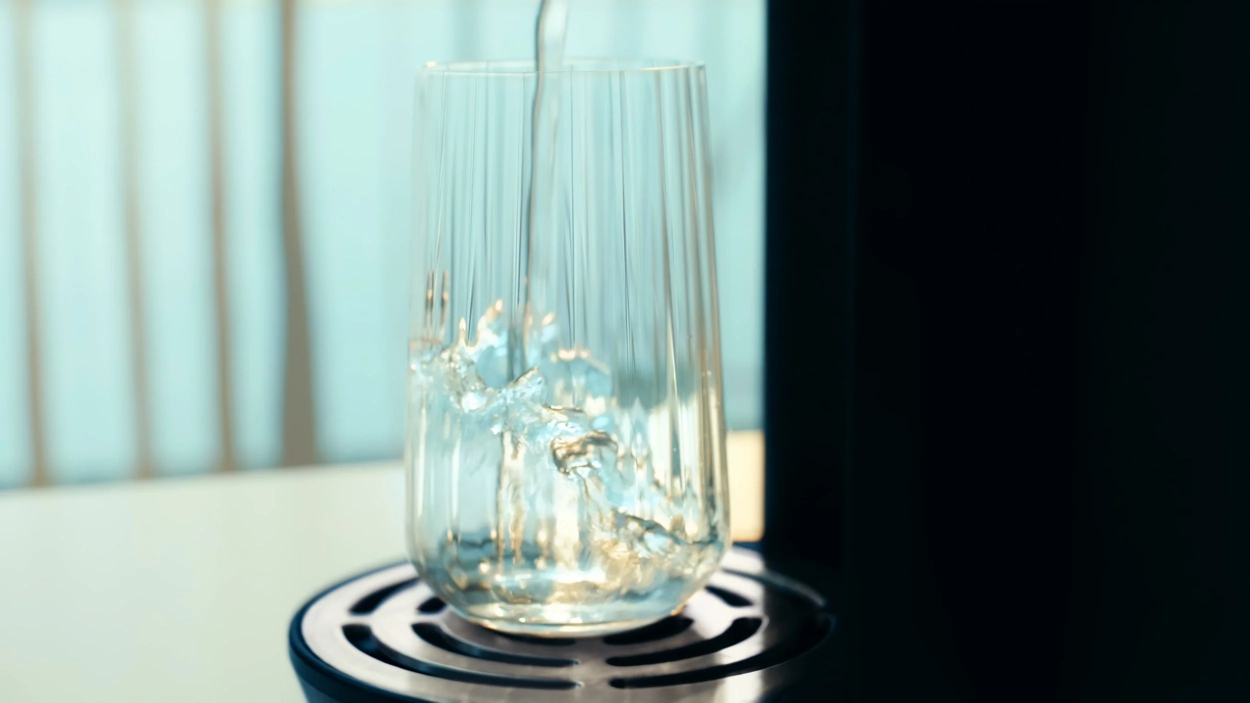A glass with water from an X2O machine being poured into it, sunlight reflecting off the water surface. The background shows a window with vertical blinds.