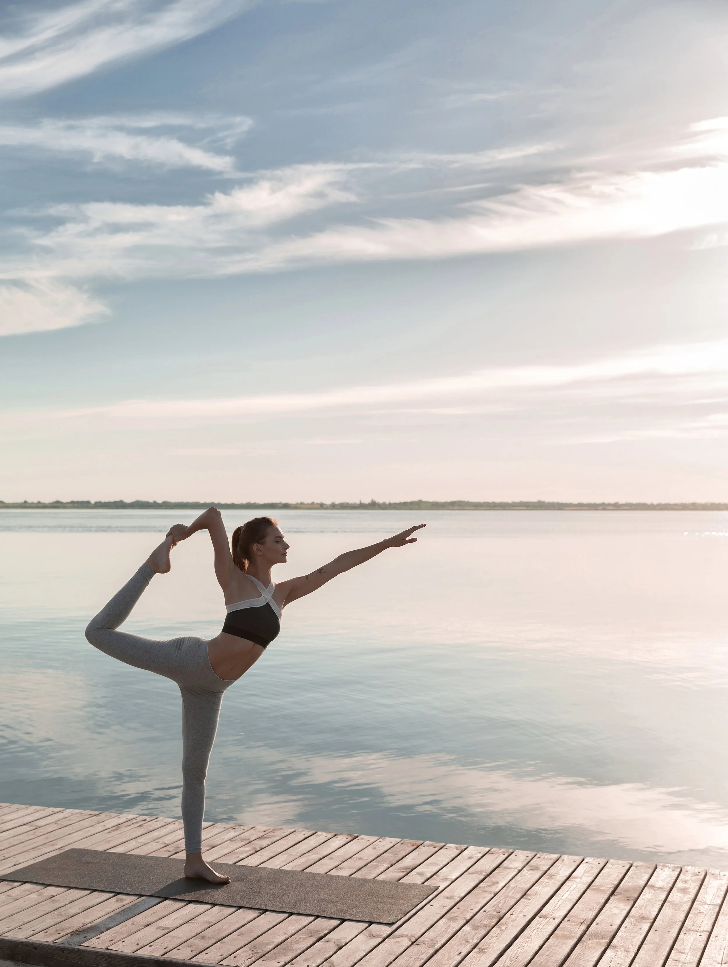 A woman practicing yoga outdoors on a wooden dock by a body of water during sunset.
