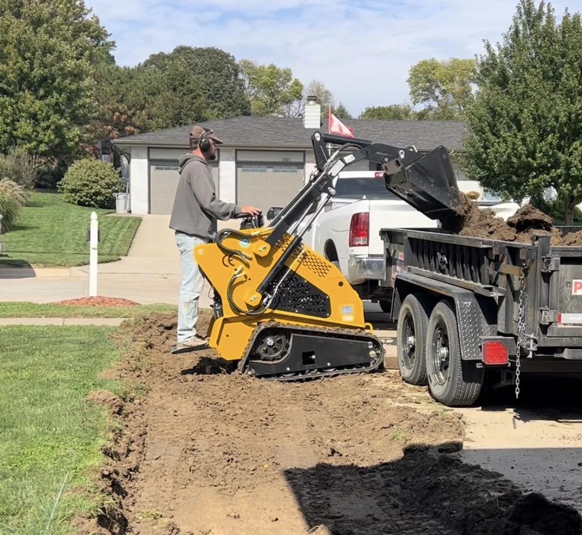 A man operating a small yellow and black excavator with tracks, loading dirt from the ground into a white dump truck on a residential street.