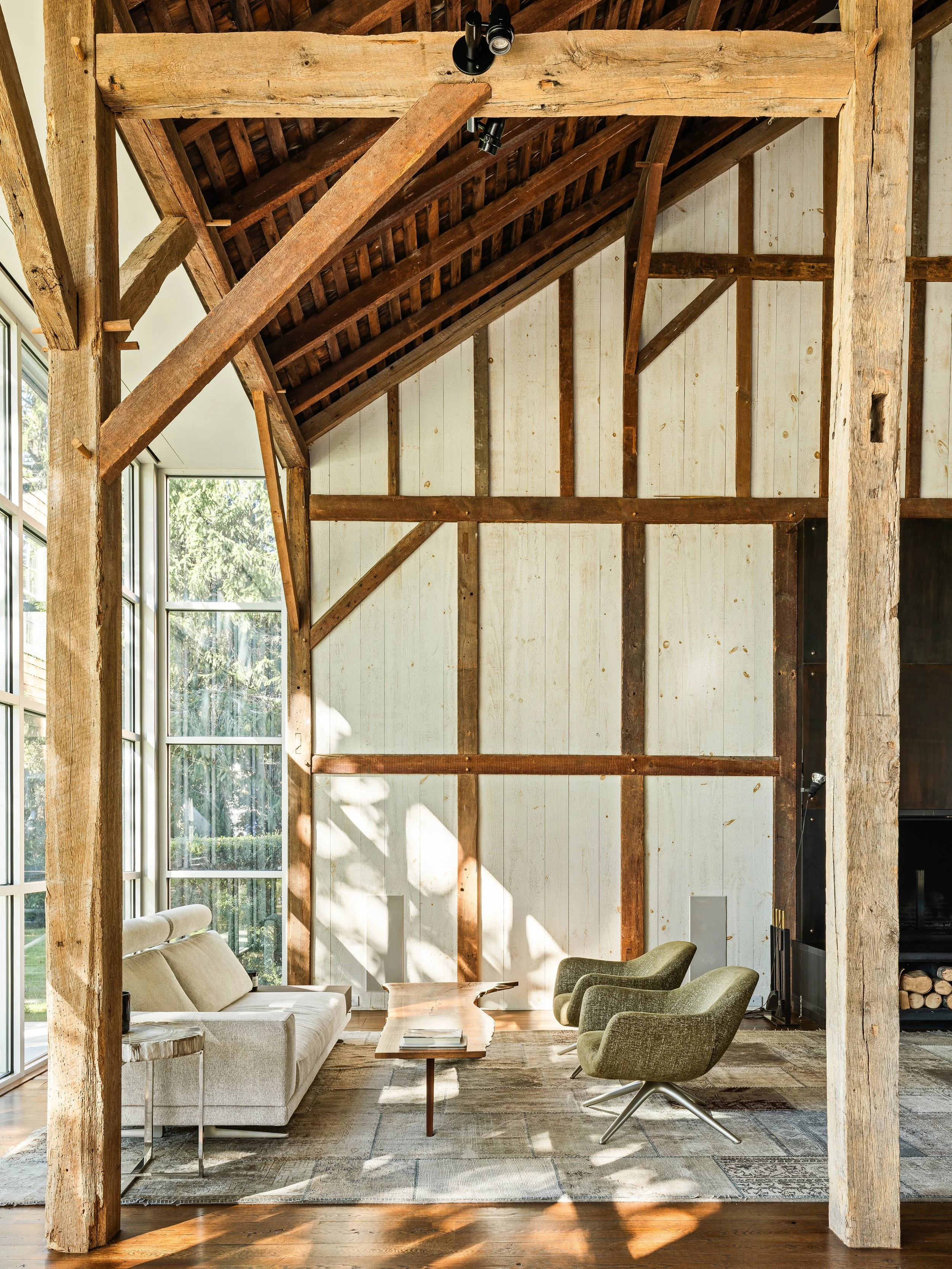 Modern living room with large windows, exposed wooden beams, a cream sofa, two green armchairs, a wooden coffee table, and a decorative rug.