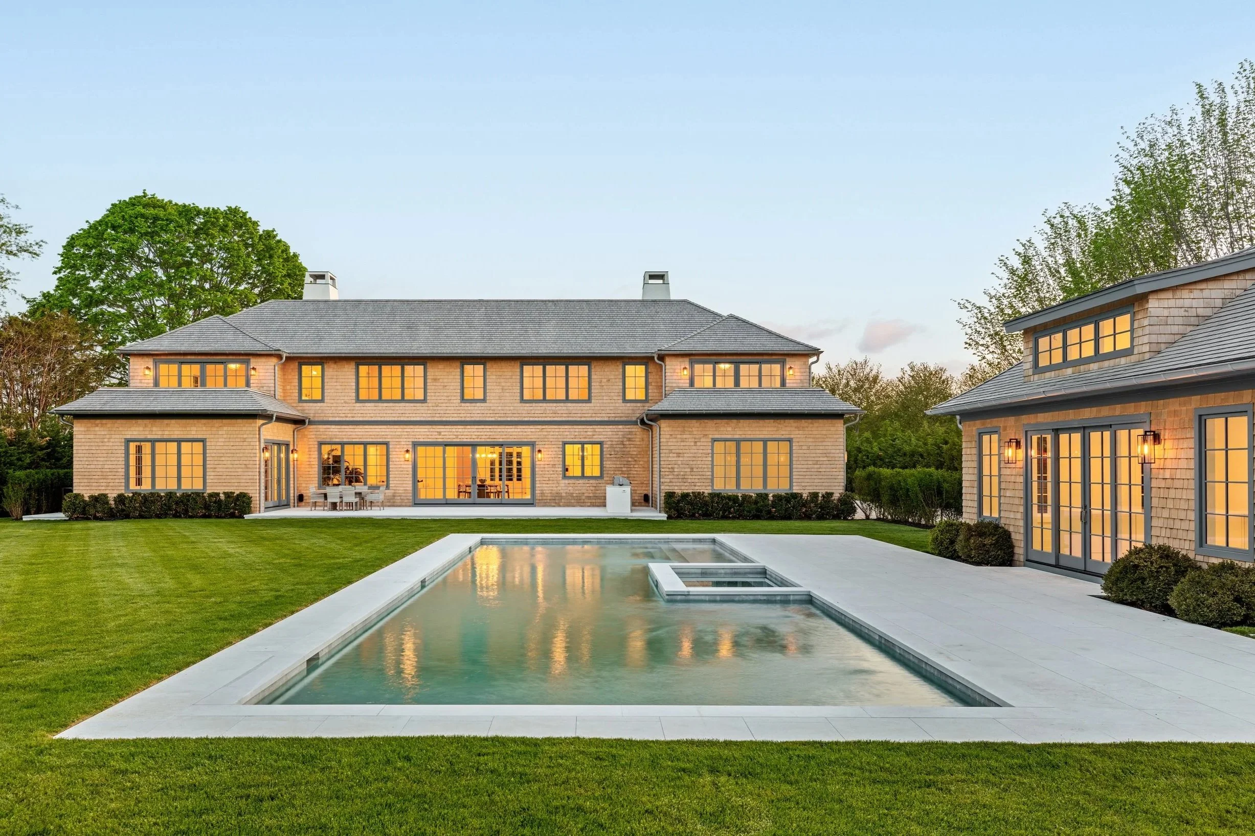 Large modern house with multiple windows and a swimming pool in the backyard, surrounded by green lawn and trees, during evening with lights on inside.