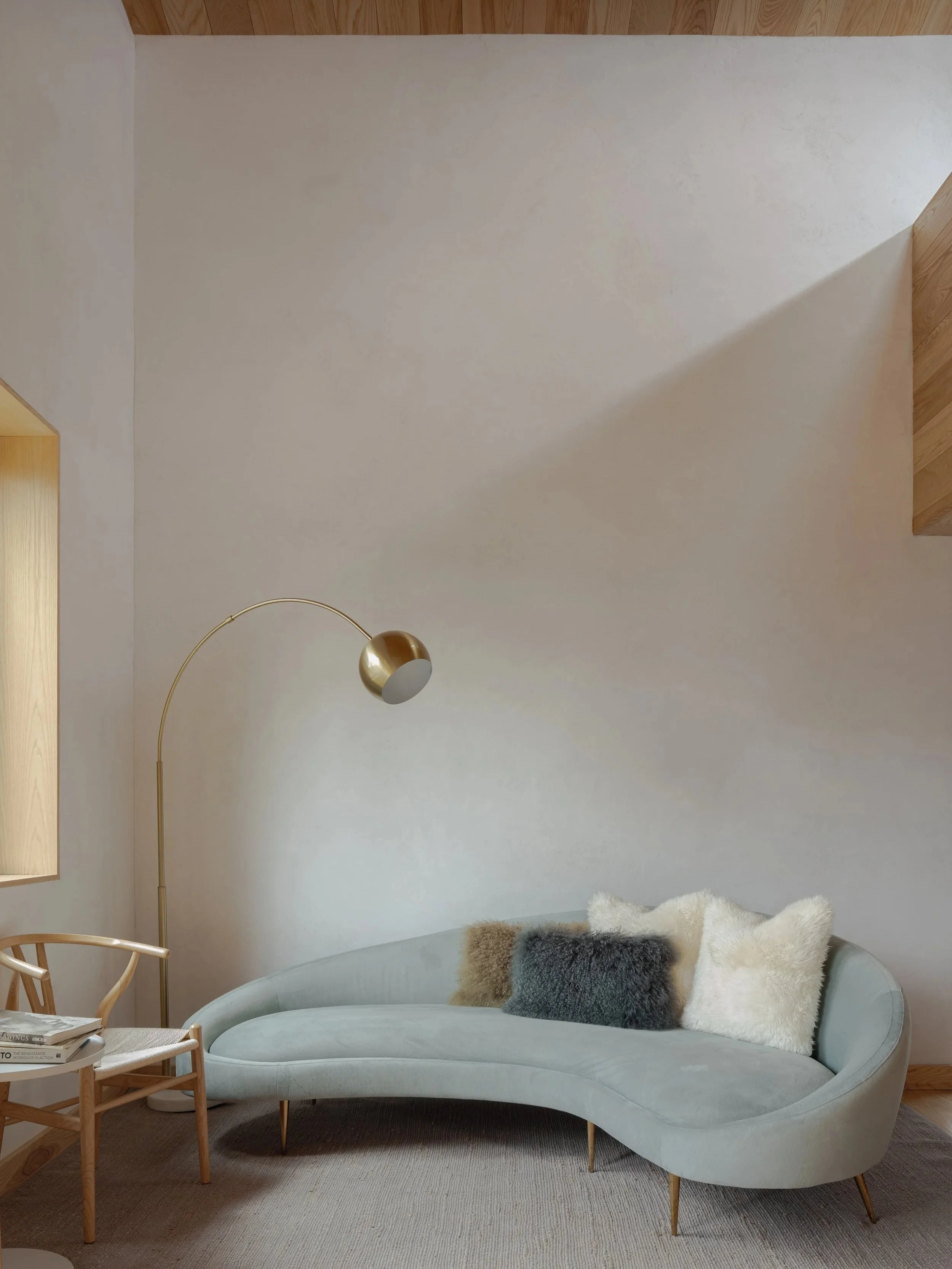 A cozy living room corner with a curved light blue velvet sofa adorned with plush fur pillows in various shades. To the left, a wooden chair with magazines on it sits next to a small side table. A modern gold floor lamp with a rounded shade arches ov