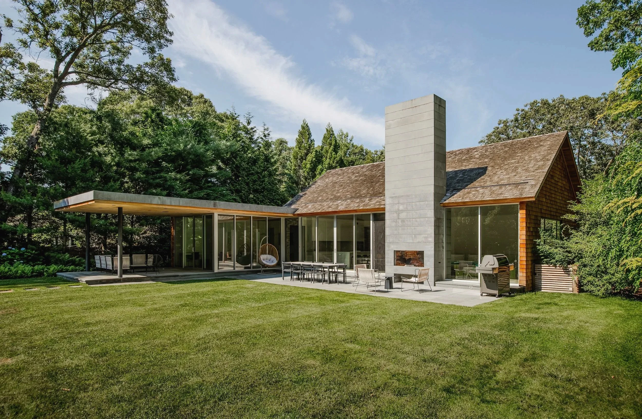 Modern house with large glass windows, a chimney, outdoor seating, and a barbecue grill, surrounded by a well-maintained lawn and trees.