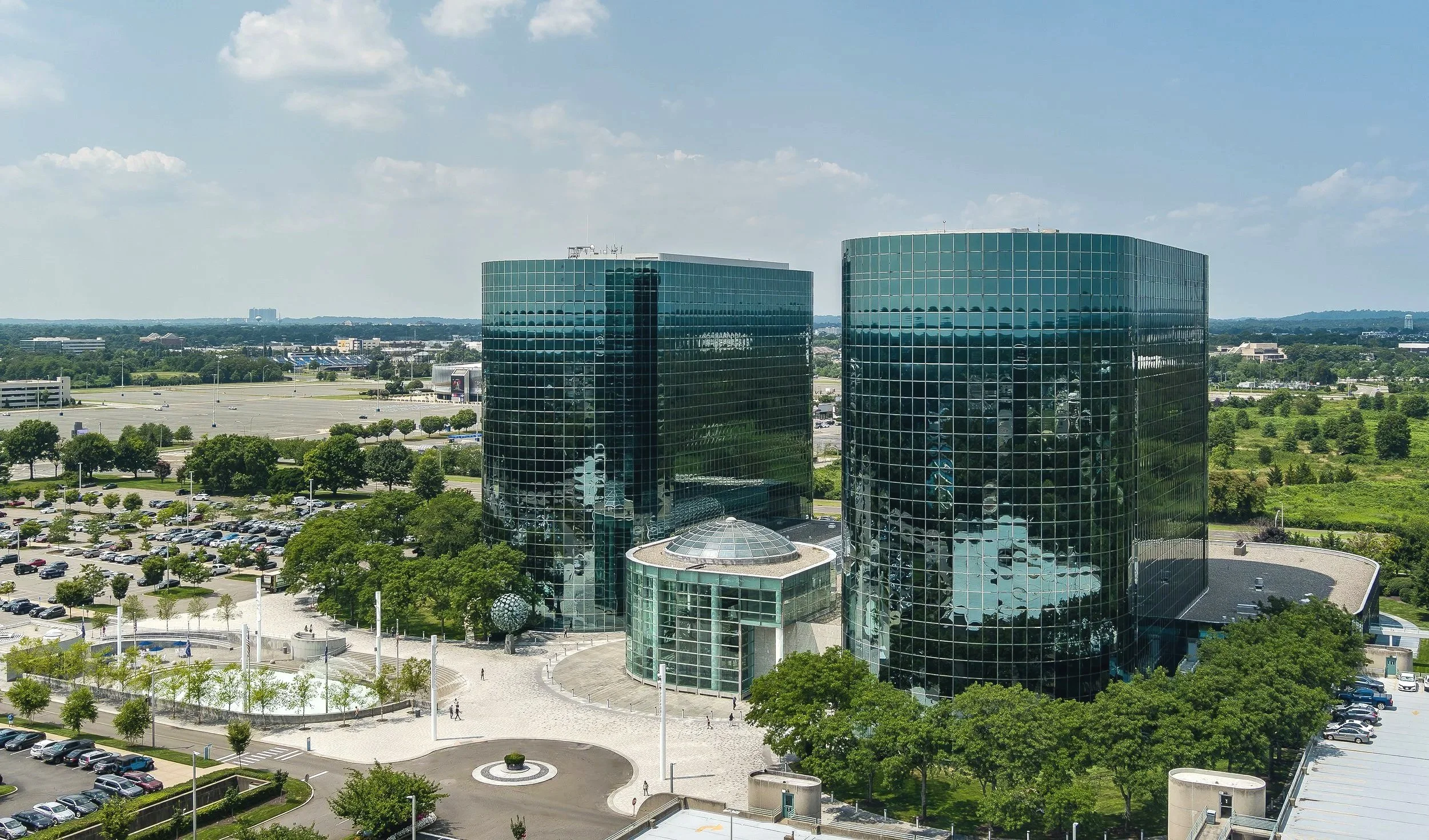 Aerial view of a modern business complex with two tall glass office buildings surrounded by trees, parking lots, and open green spaces under a blue sky with scattered clouds.