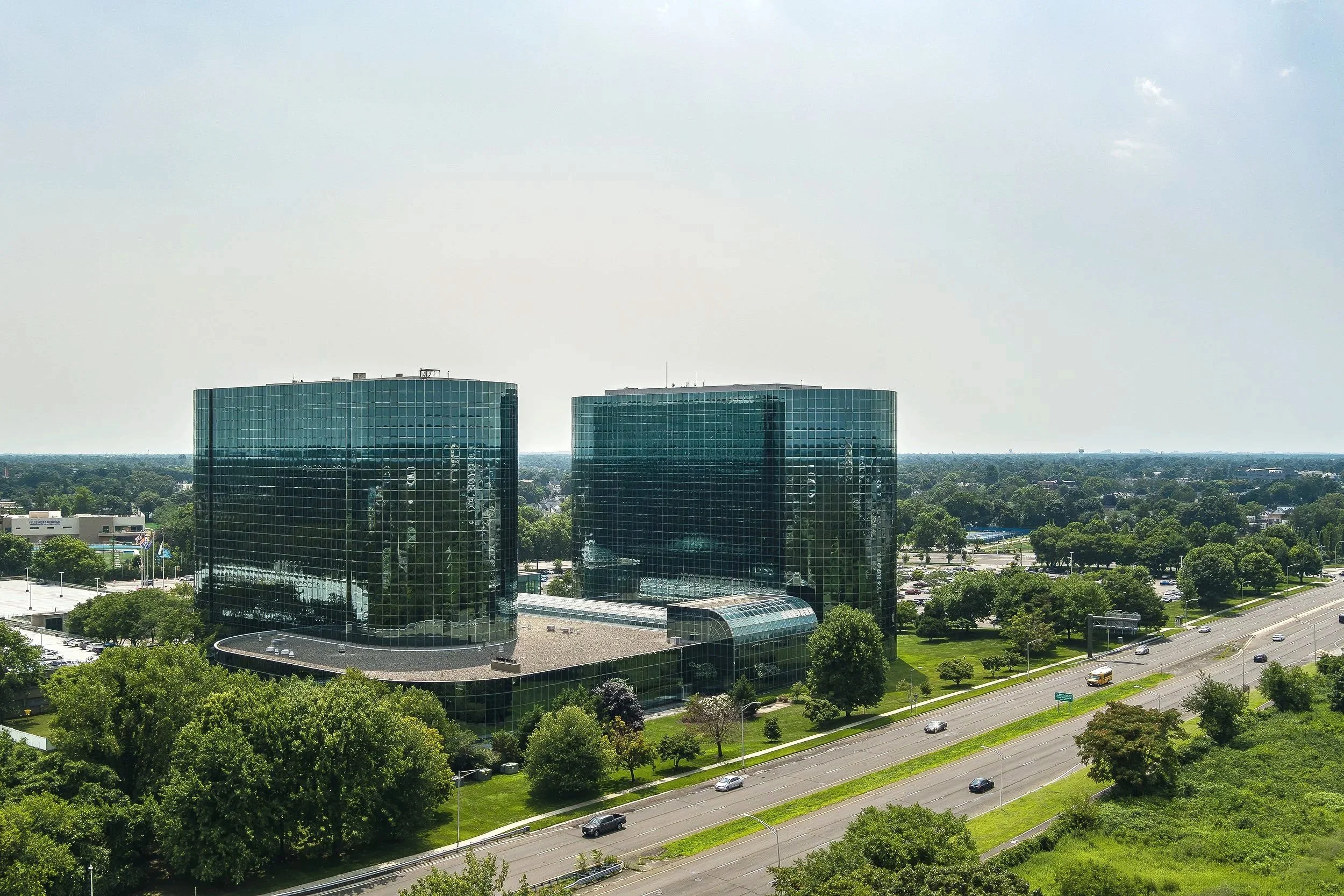 Two tall, black glass office buildings with curved sides and reflective surfaces, set in a green landscape with trees, grass, and a road with passing cars.