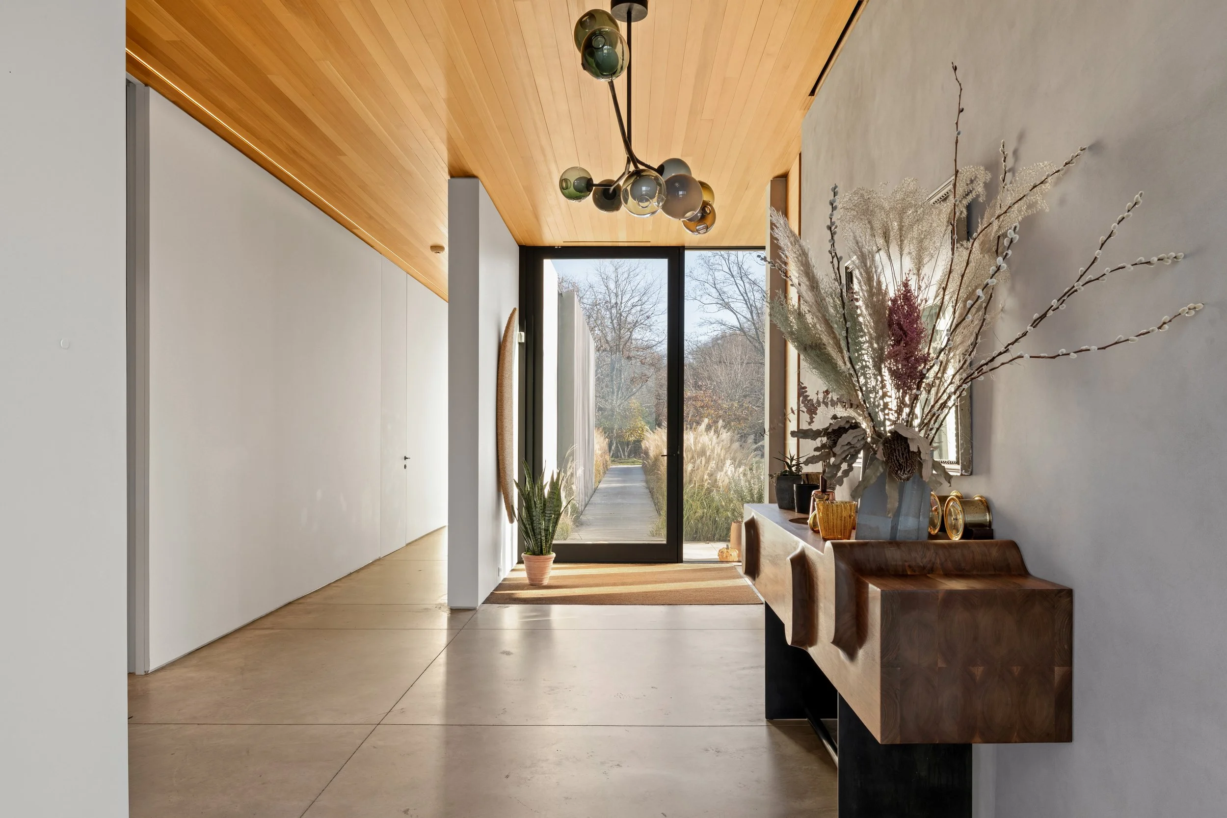 Entryway with wooden ceiling, modern chandelier, glass front door, indoor plant, and decorative sideboard with large dried flower arrangement.