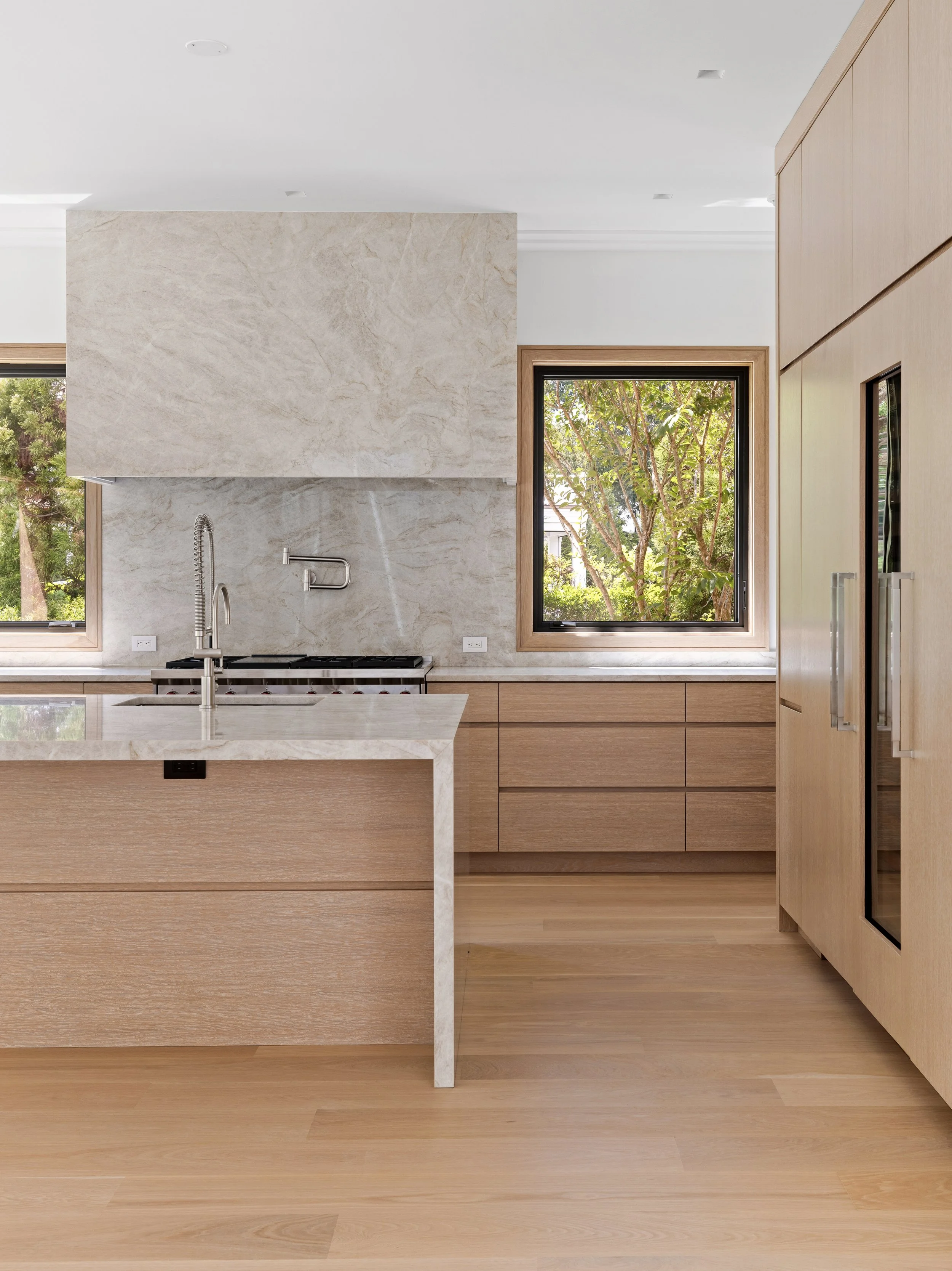Modern kitchen with light wood cabinets, marble countertops, a sink, and large windows showing trees outside.