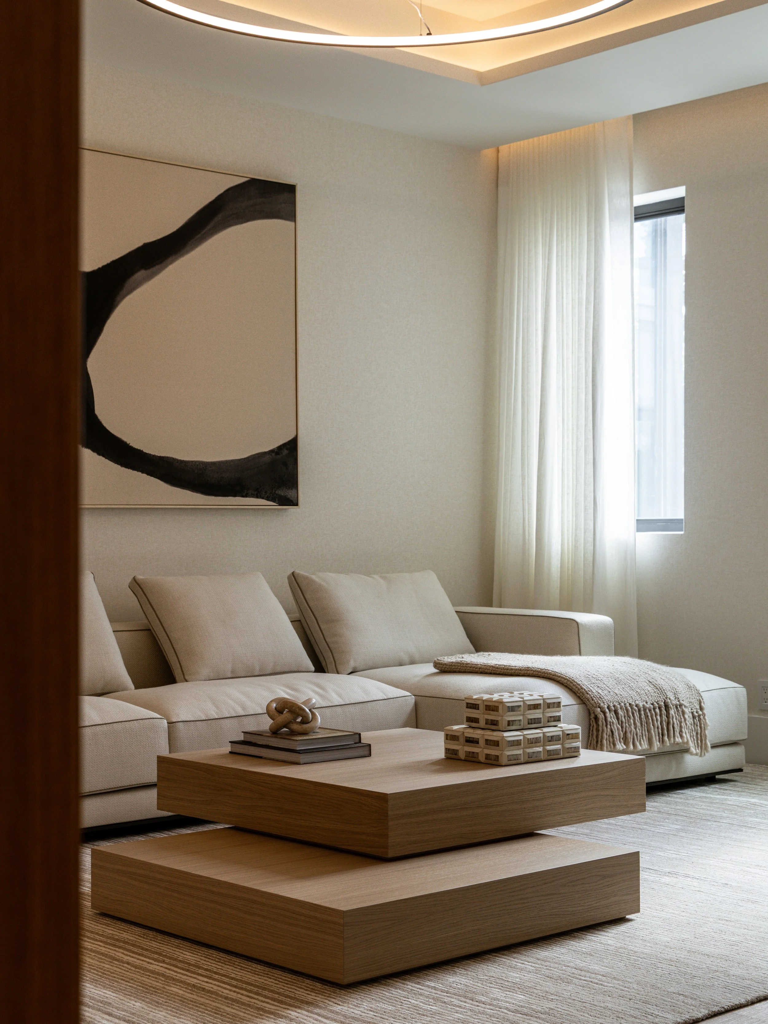 Modern living room with beige sofa, wooden coffee table, abstract wall art, and window with sheer curtains