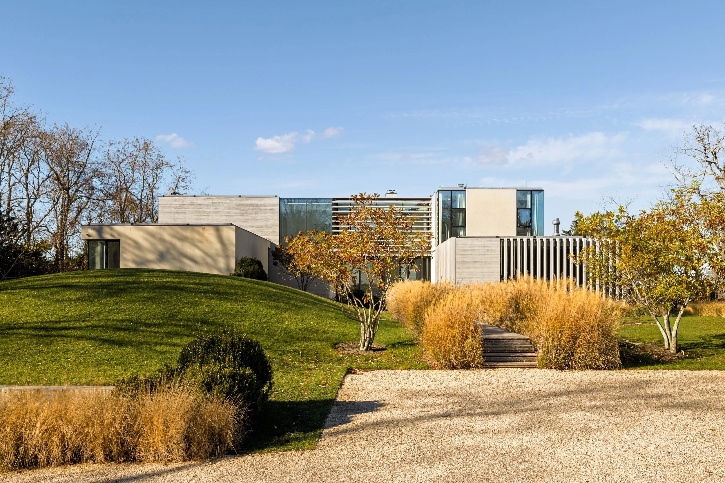 Modern house with beige walls and large glass windows on a landscaped lawn with trees, shrubs, and walking paths under a blue sky.