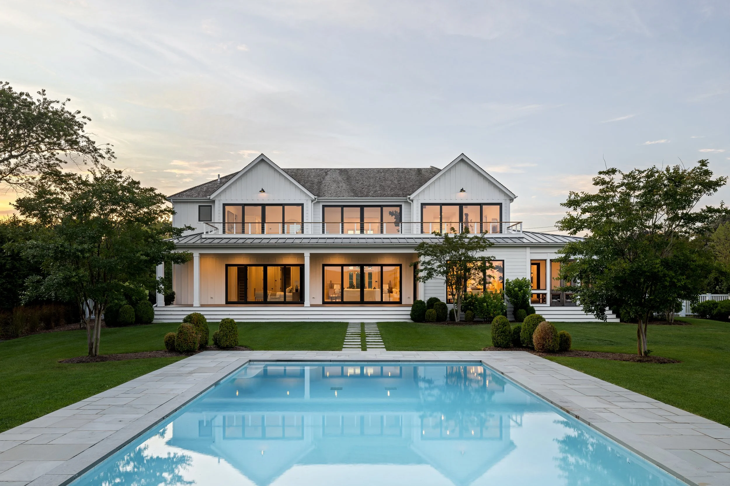 A white two-story house with large glass windows and a balcony, surrounded by a lush green lawn, with a swimming pool in the foreground and trees on either side, during sunset