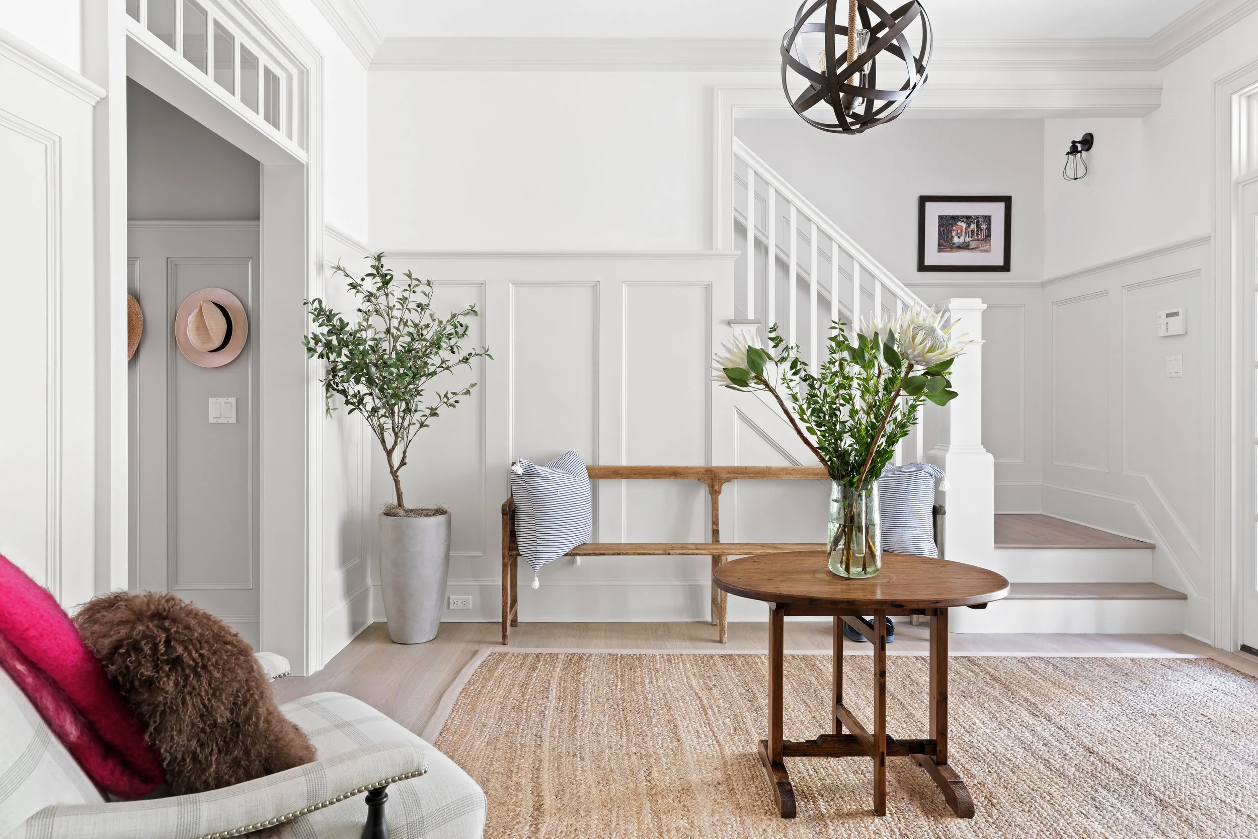 Bright, modern entryway with white walls, wooden staircase, a small wooden table with a vase of flowers, a bench with striped pillows, a potted tree, and decorative hats on the wall.