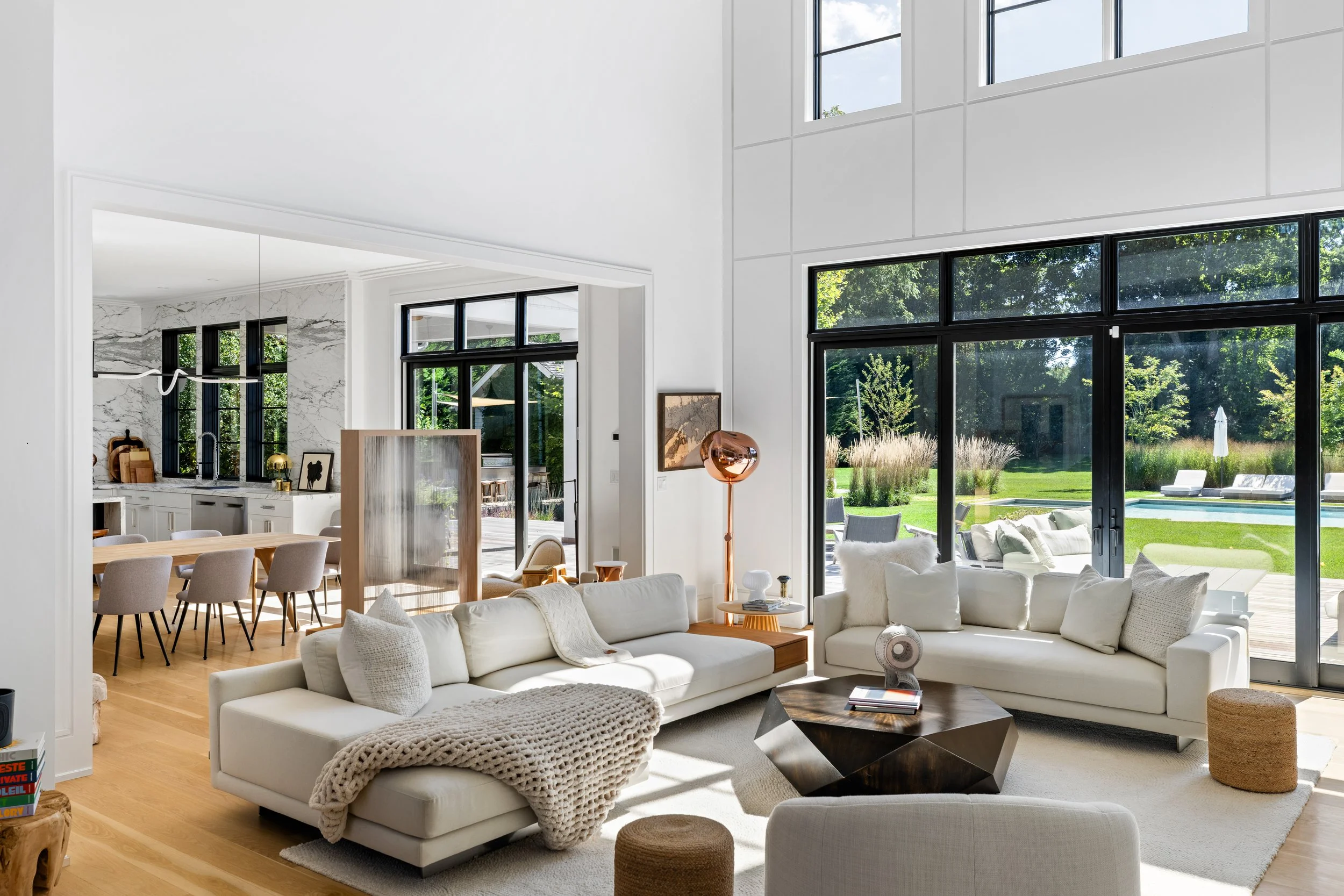 Bright living room with white L-shaped sectional sofa, beige pillows, and cozy knit blanket, large glass windows showing lush green outdoor scenery, wooden coffee table with decorative objects, in a modern open-plan house with minimalistic decor.