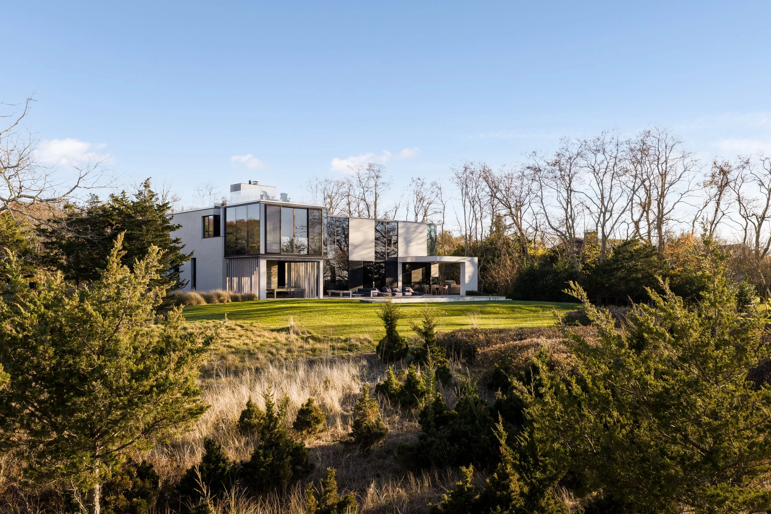 Modern house with large glass windows on a grassy hill, surrounded by trees and shrubs, under a blue sky.
