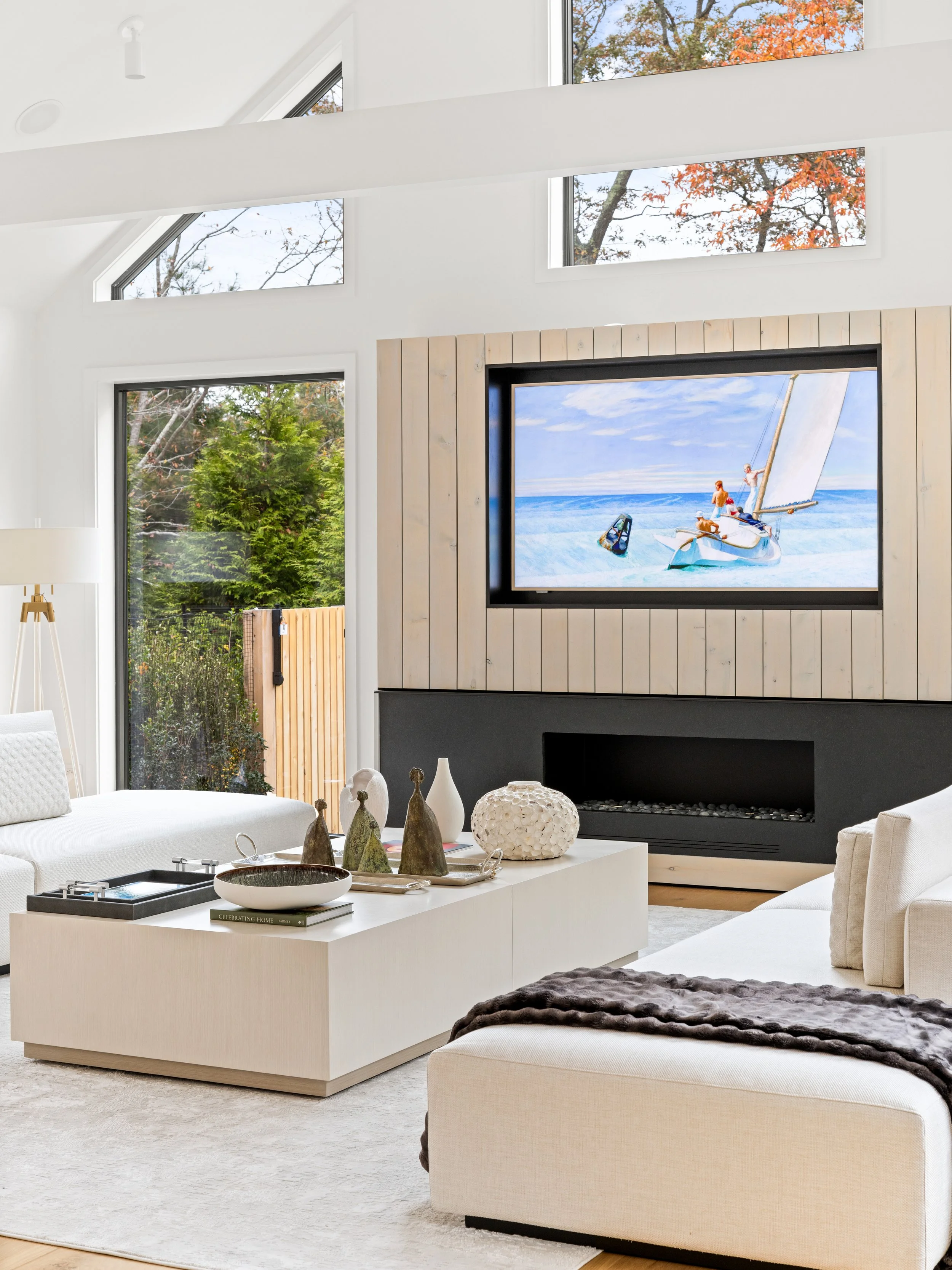 Modern living room with white walls, large windows, a black fireplace, a wall-mounted television displaying a sailing scene, and white furniture with decorative objects on the coffee table.