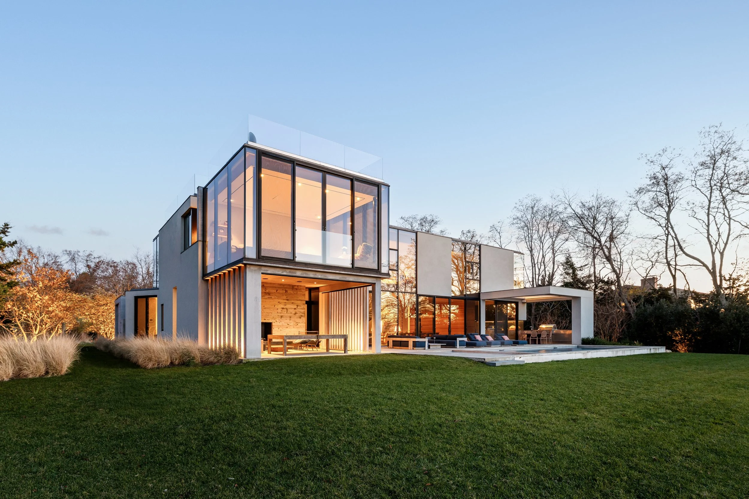 Modern house with large glass windows, outdoor seating, and a well-maintained lawn under a clear sky at sunset.