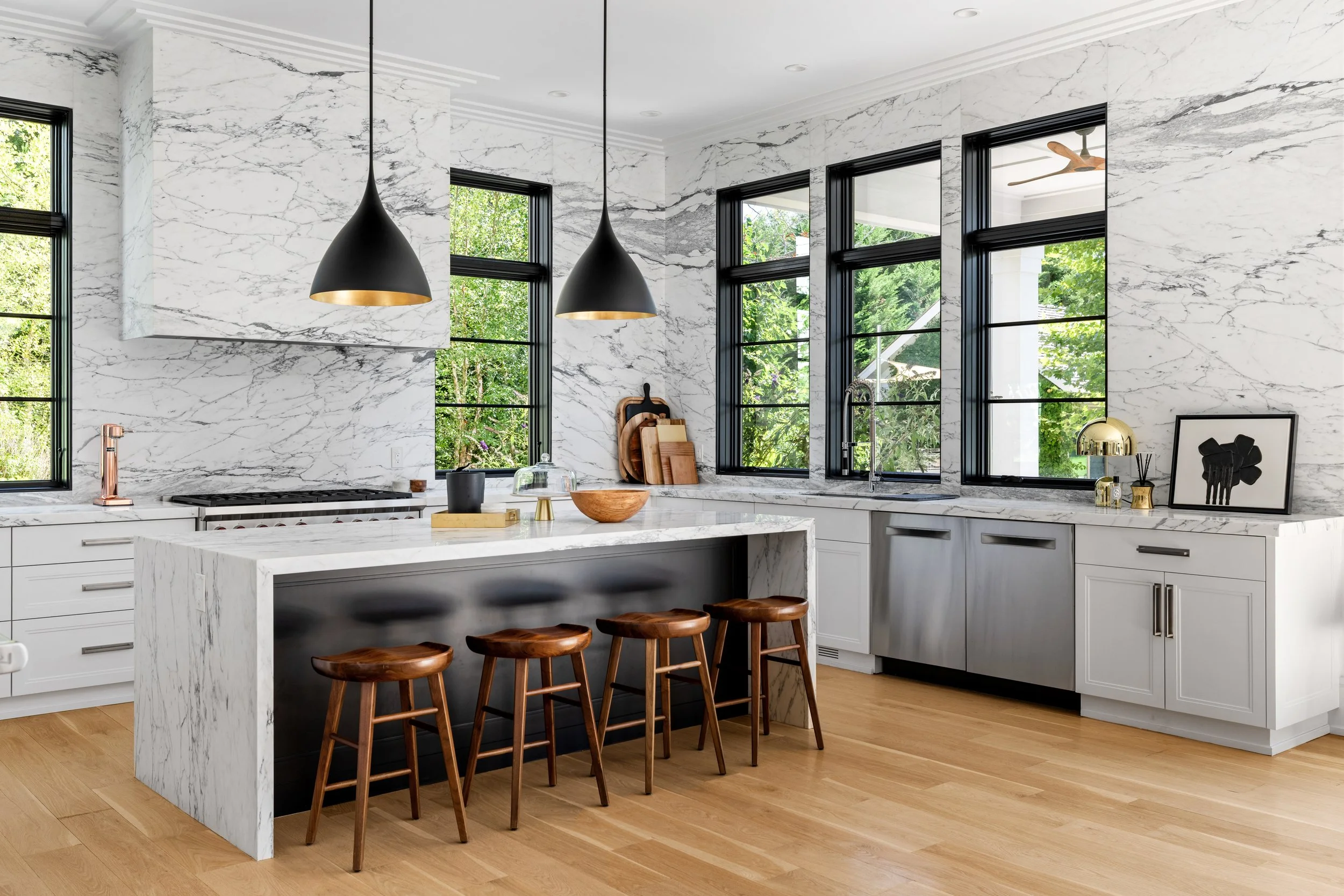 Modern kitchen with white marble walls, black window frames, a marble island with wooden bar stools, and black pendant lights. There are kitchen tools and decor on the countertops, with a view of greenery outside.