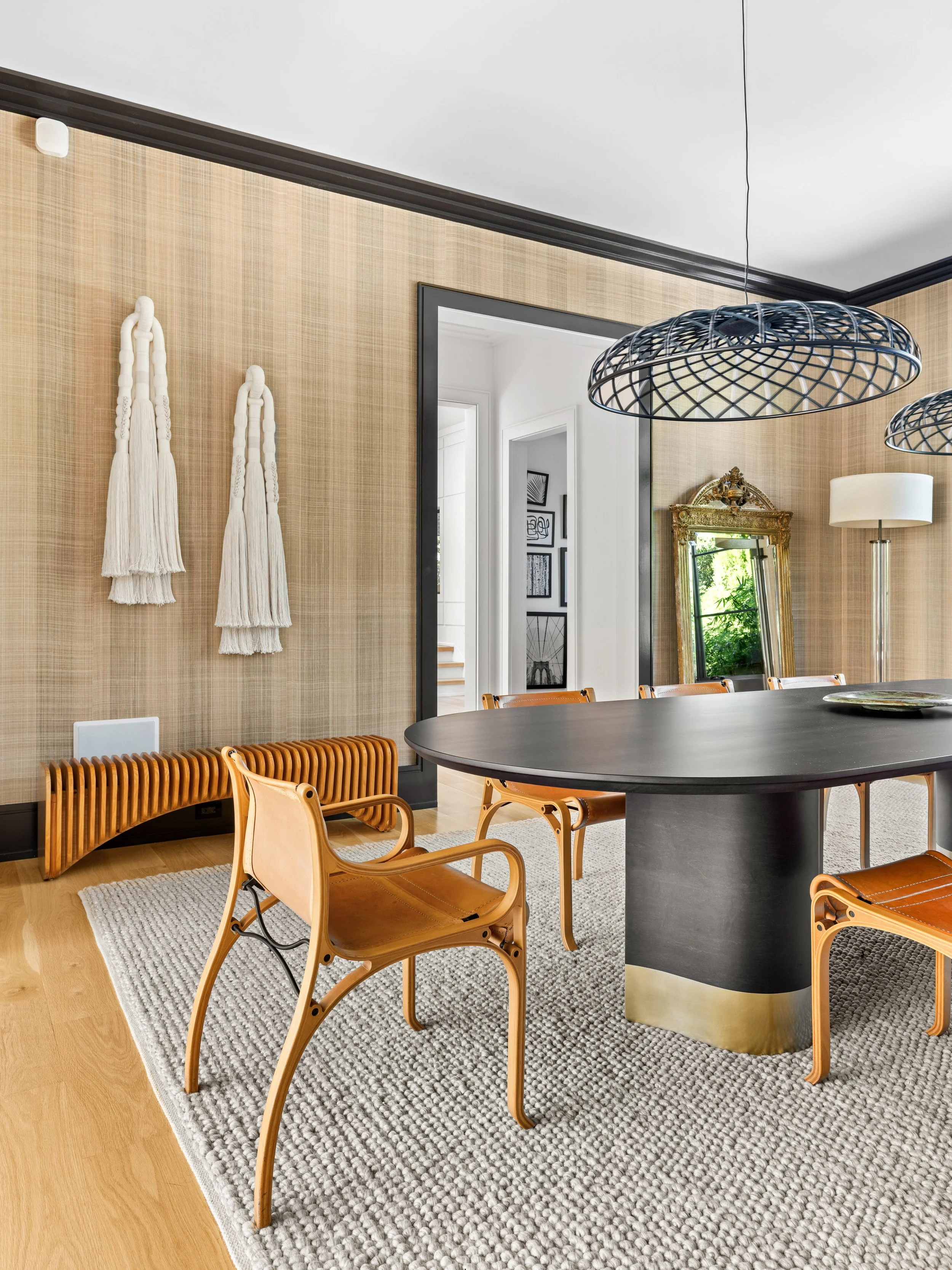 Modern dining room with a black oval table, wooden chairs, beige textured wall, white tassel wall hangings, gold-trimmed mirror, wicker light fixtures, and a tall floor lamp.