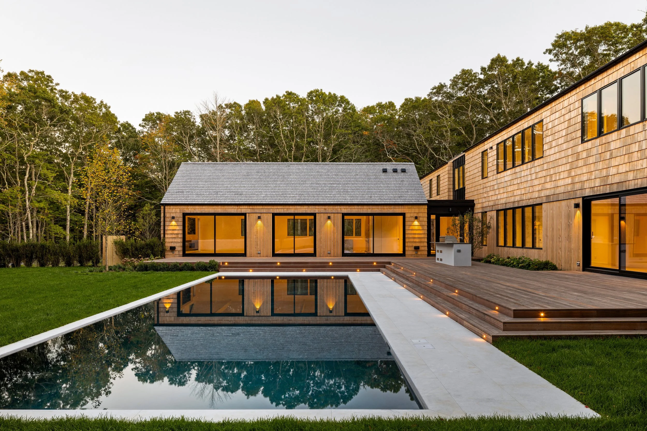 Modern house with large glass doors, wooden exterior, surrounded by a well-manicured lawn, with a rectangular pool reflecting the house and trees, and a wooded background.
