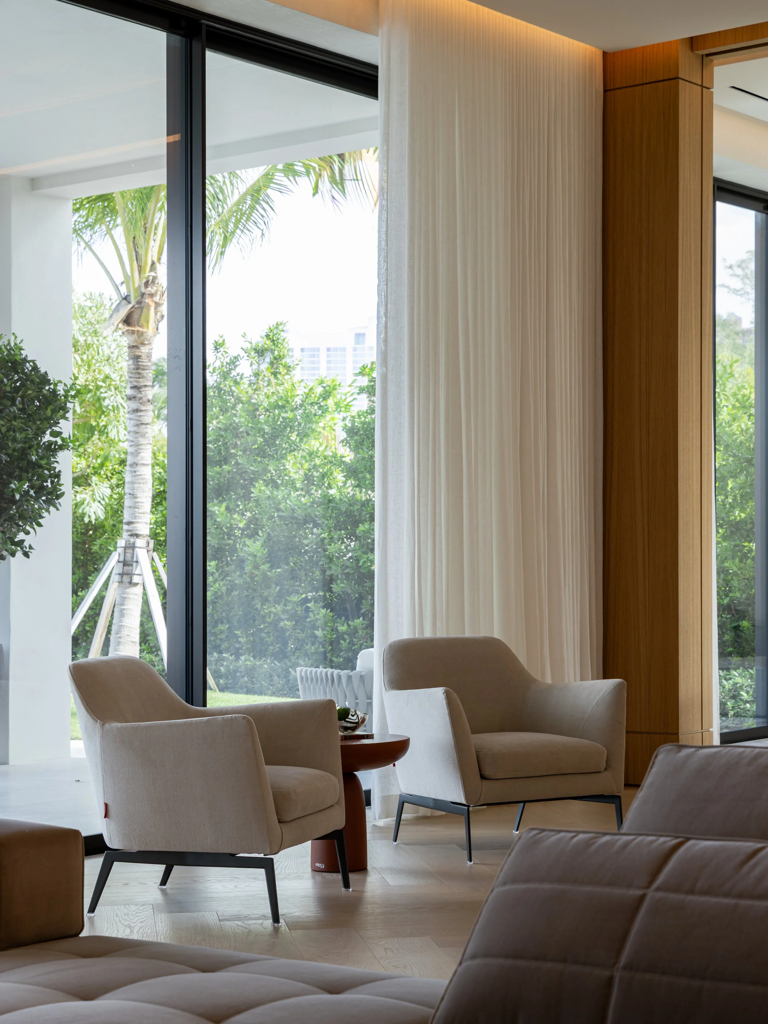 A modern living room with beige armchairs, a small round wooden table, large floor-to-ceiling windows with white curtains, and lush greenery outside including a palm tree.