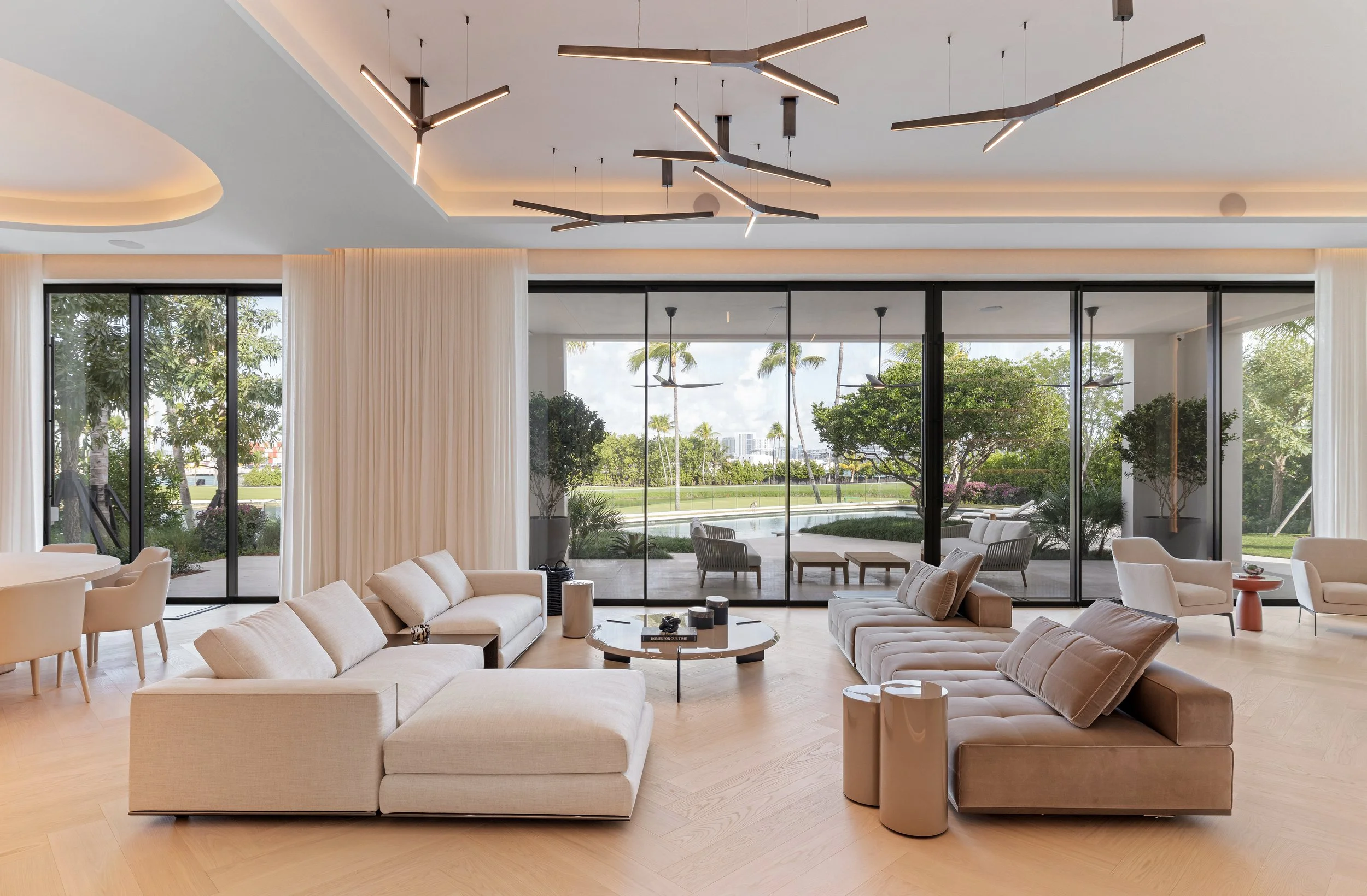 Modern living room with large glass sliding doors showing an outdoor pool and landscaped garden, beige sectional sofas, a round coffee table, contemporary lighting fixtures, and neutral-toned decor.