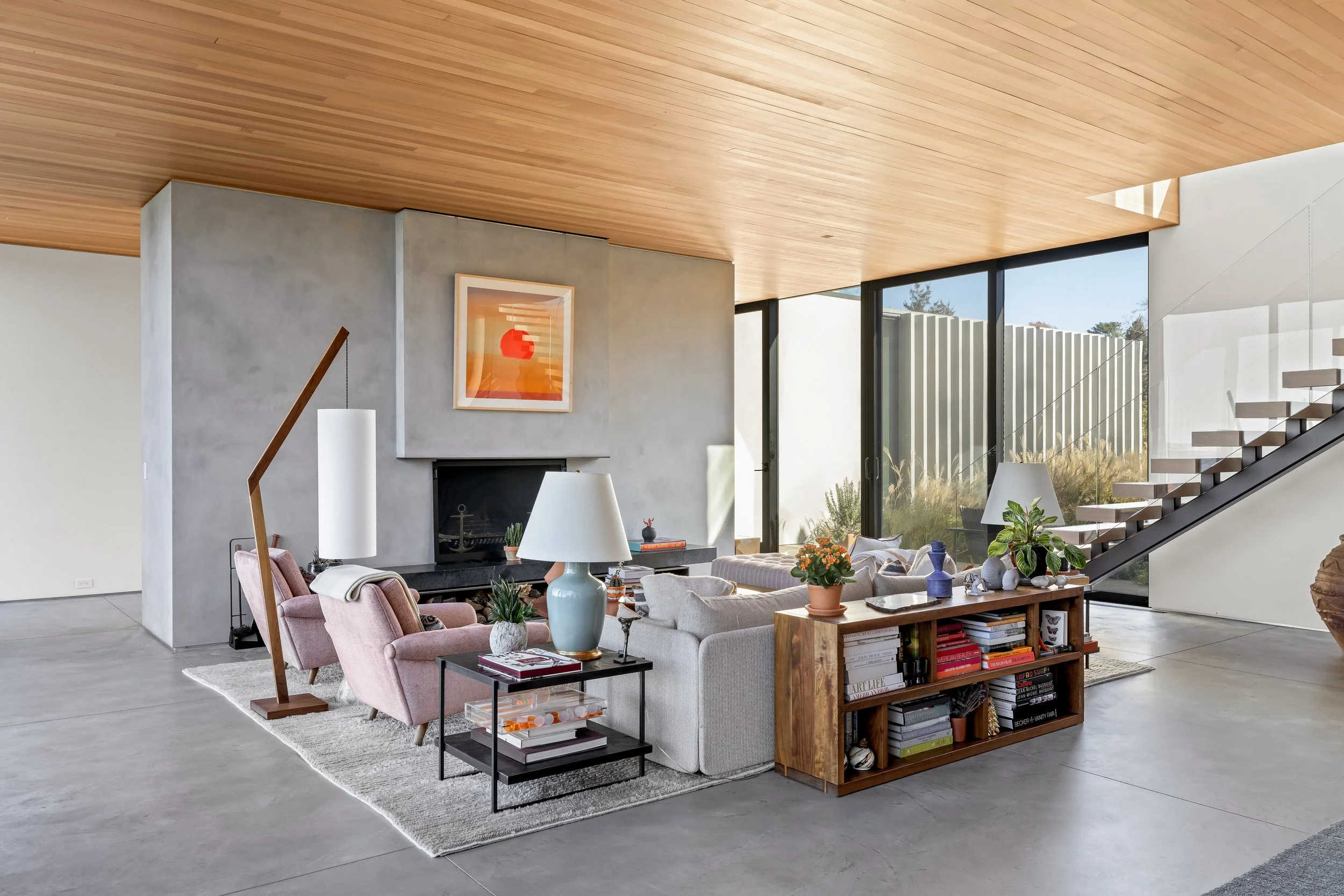 Modern living room with concrete floors, wooden ceiling, and large glass windows showing outdoors. Features a fireplace, gray and pink sofas, a wooden bookshelf, and various decorative items.