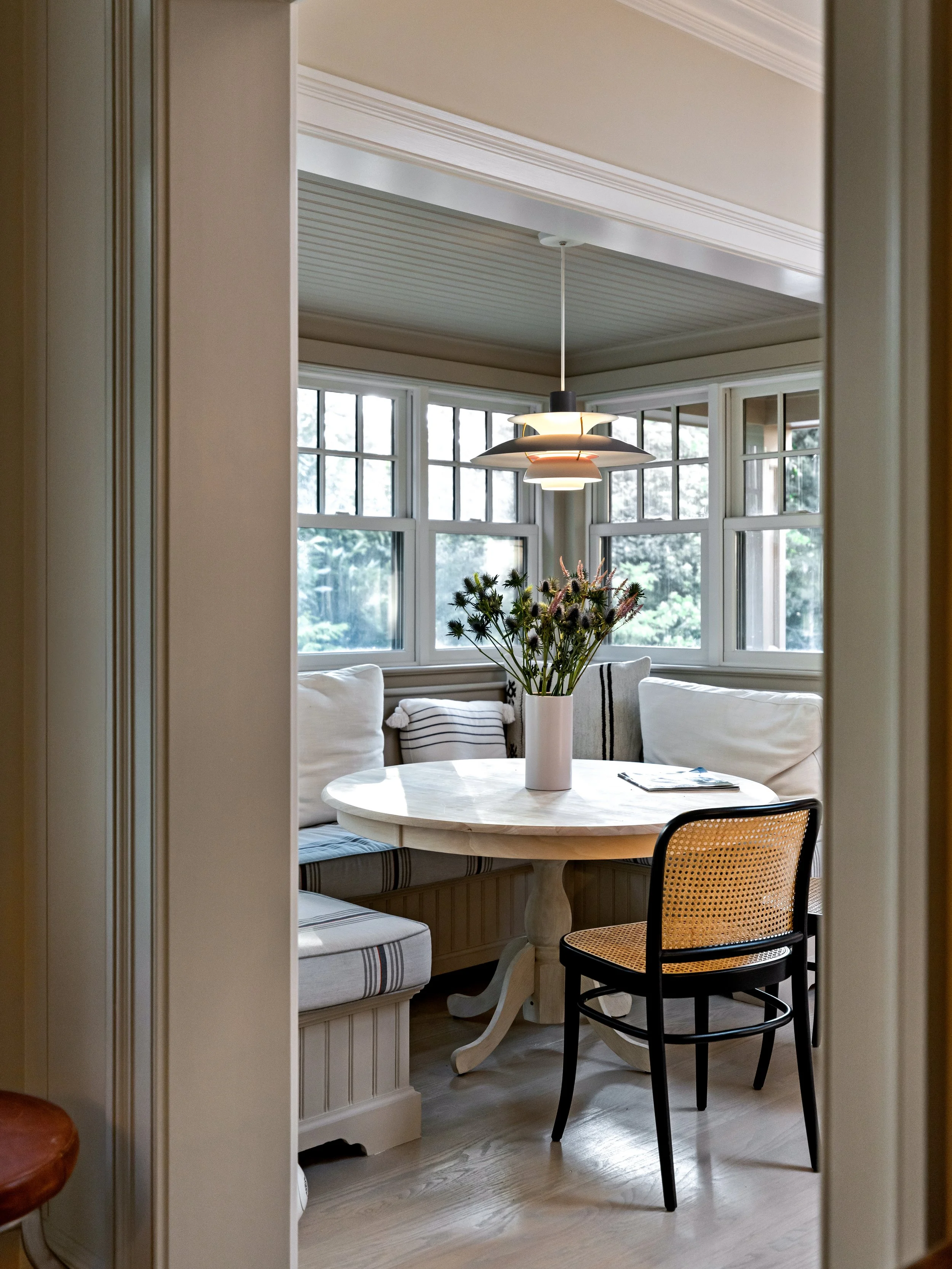 A cozy dining nook with a round marble table, surrounded by white cushioned benches and a black chair with a rattan seat, decorated with a vase of flowers and a small stack of magazines, inside a bright room with large windows.