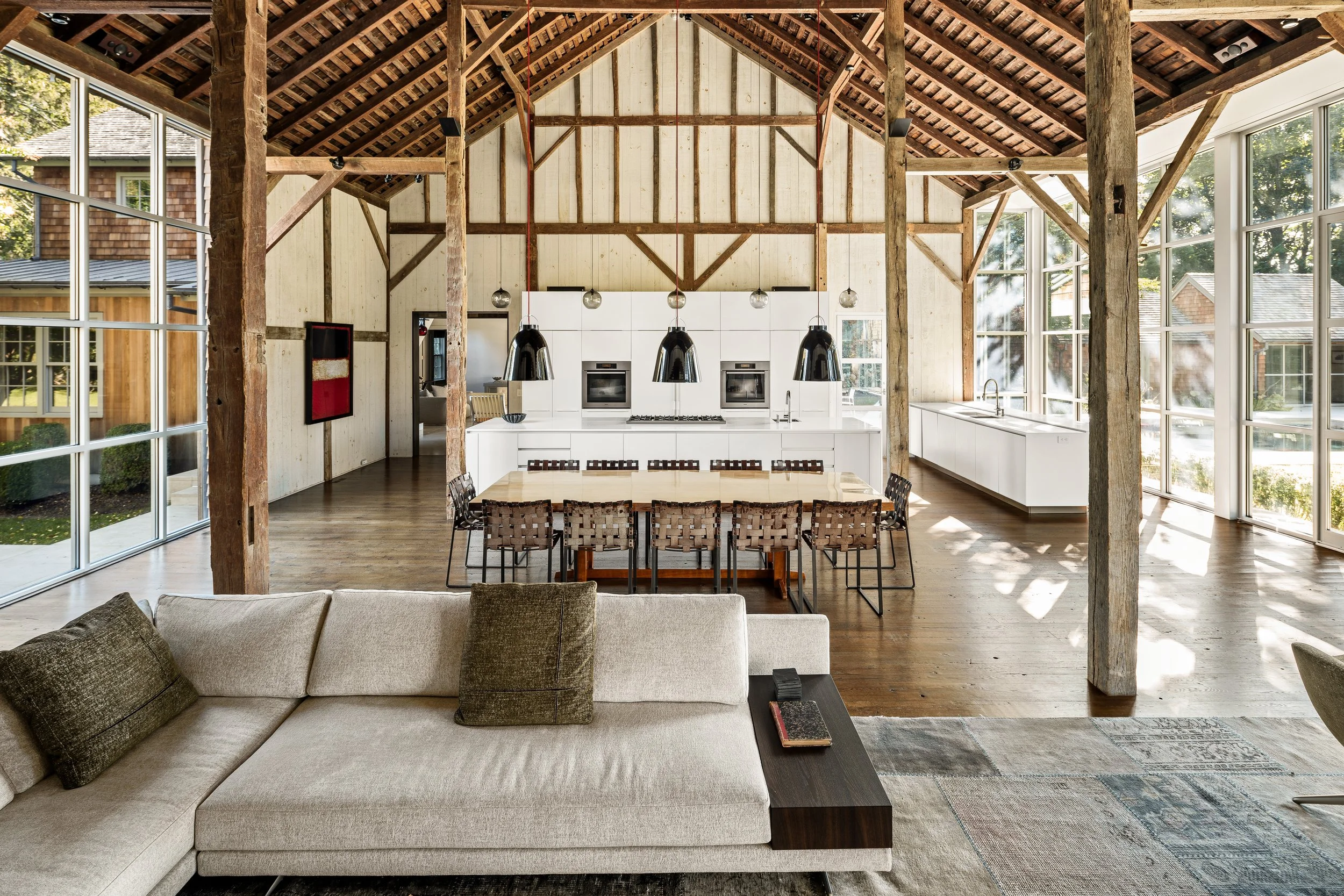 Interior of a modern open-concept living space with large windows, rustic wooden beams, a white kitchen with built-in ovens, a luxurious dining table, and a beige sofa in the foreground.