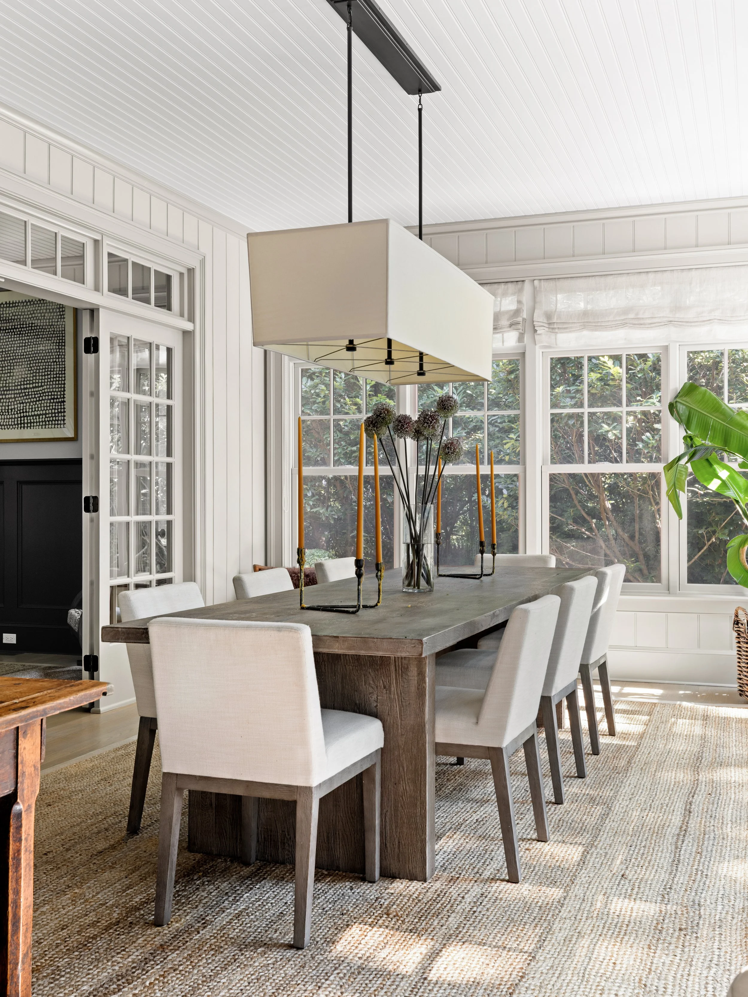 A dining room with a long wooden table, nine white upholstered chairs, a large rectangular pendant light, and a glass vase with tall flowers, surrounded by windows with white framing, greenery outside, and a glass door to the left.