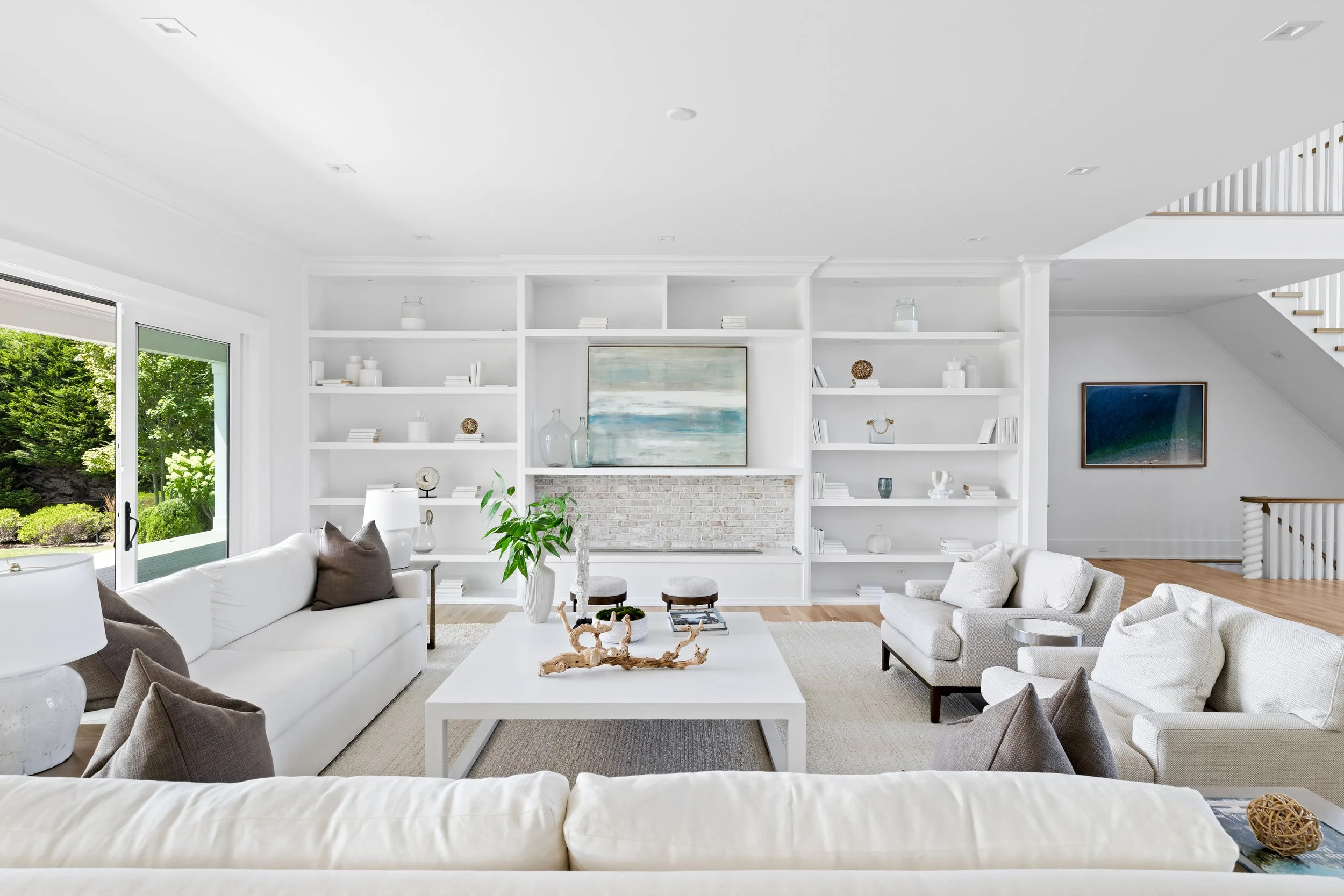 Bright living room with white sofas, a large window showing green outdoor scenery, white built-in shelves with decor, and a staircase on the right.
