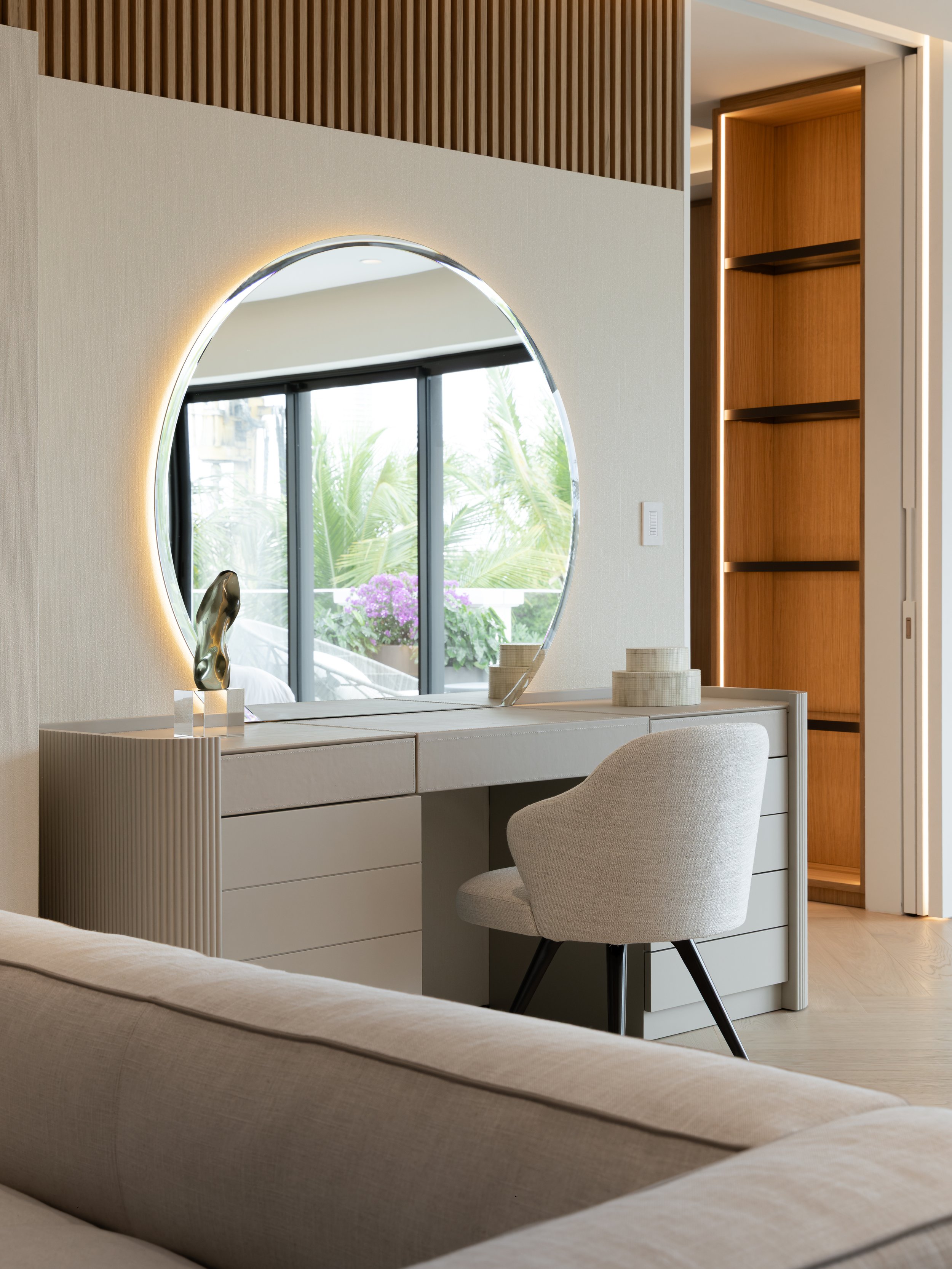 Modern bedroom with a beige desk and a white upholstered chair, a large round mirror with backlighting, and a sliding glass door with greenery outside.
