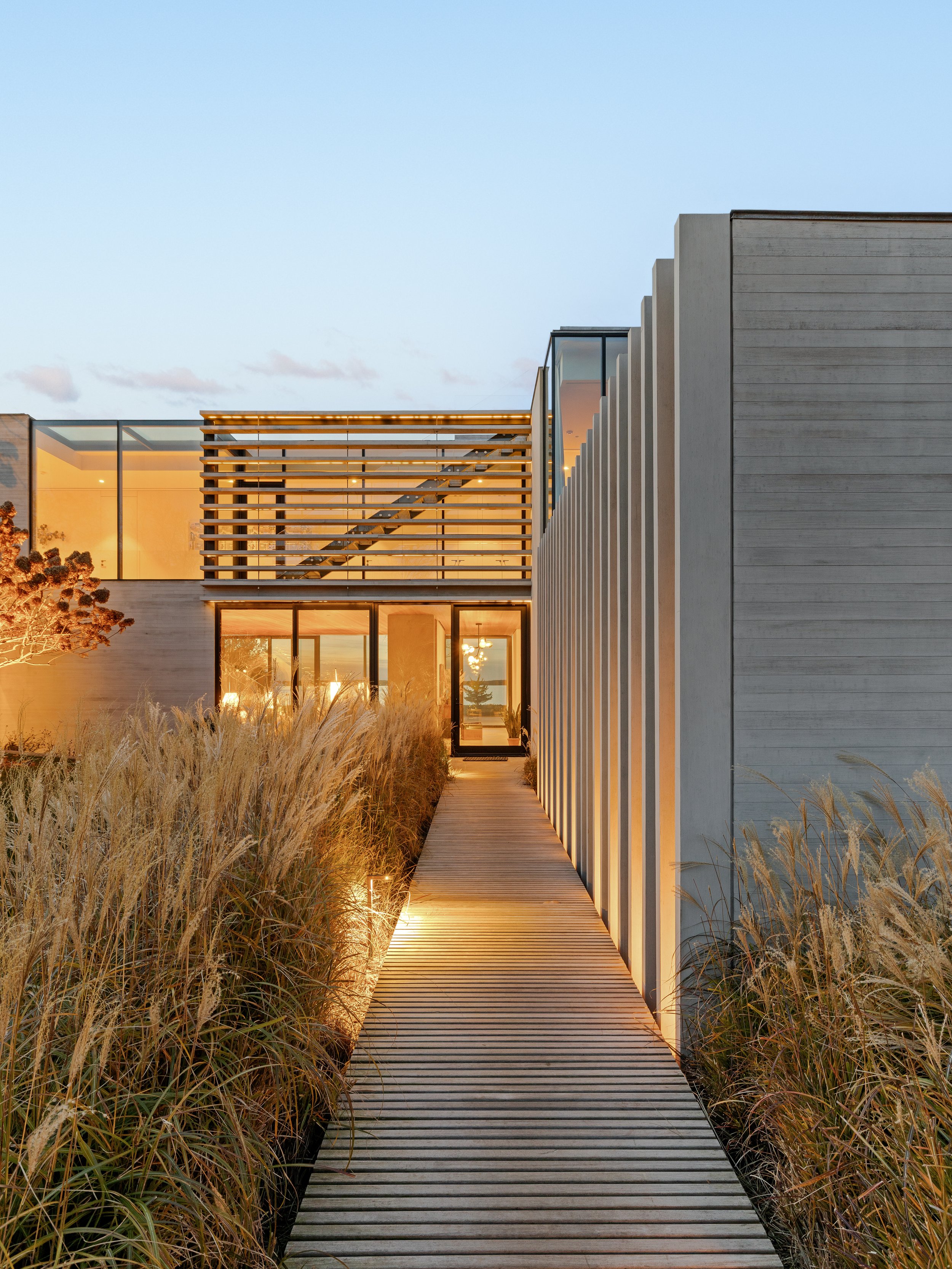 A modern house with large glass windows, a wood walkway leading to the entrance, and a landscaped yard with tall grasses, illuminated by outdoor lighting during sunset or dusk.