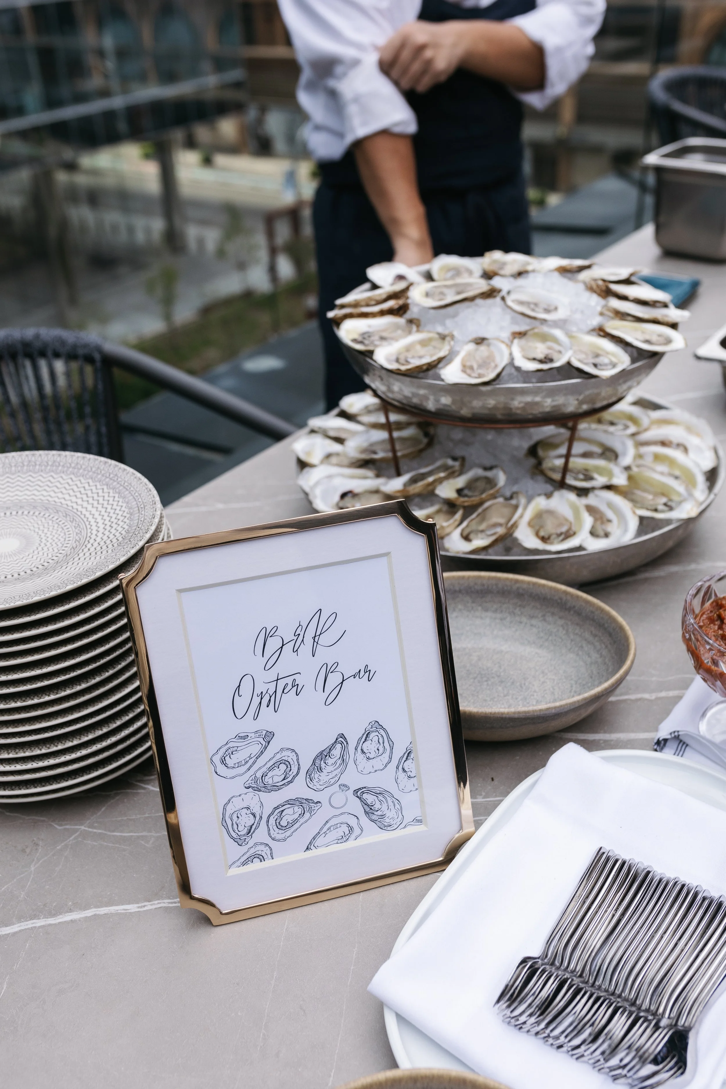 A table with a sign reading 'B&L Oyster Bar,' stacks of plates, a tray of oysters on ice, and a server standing behind the table.