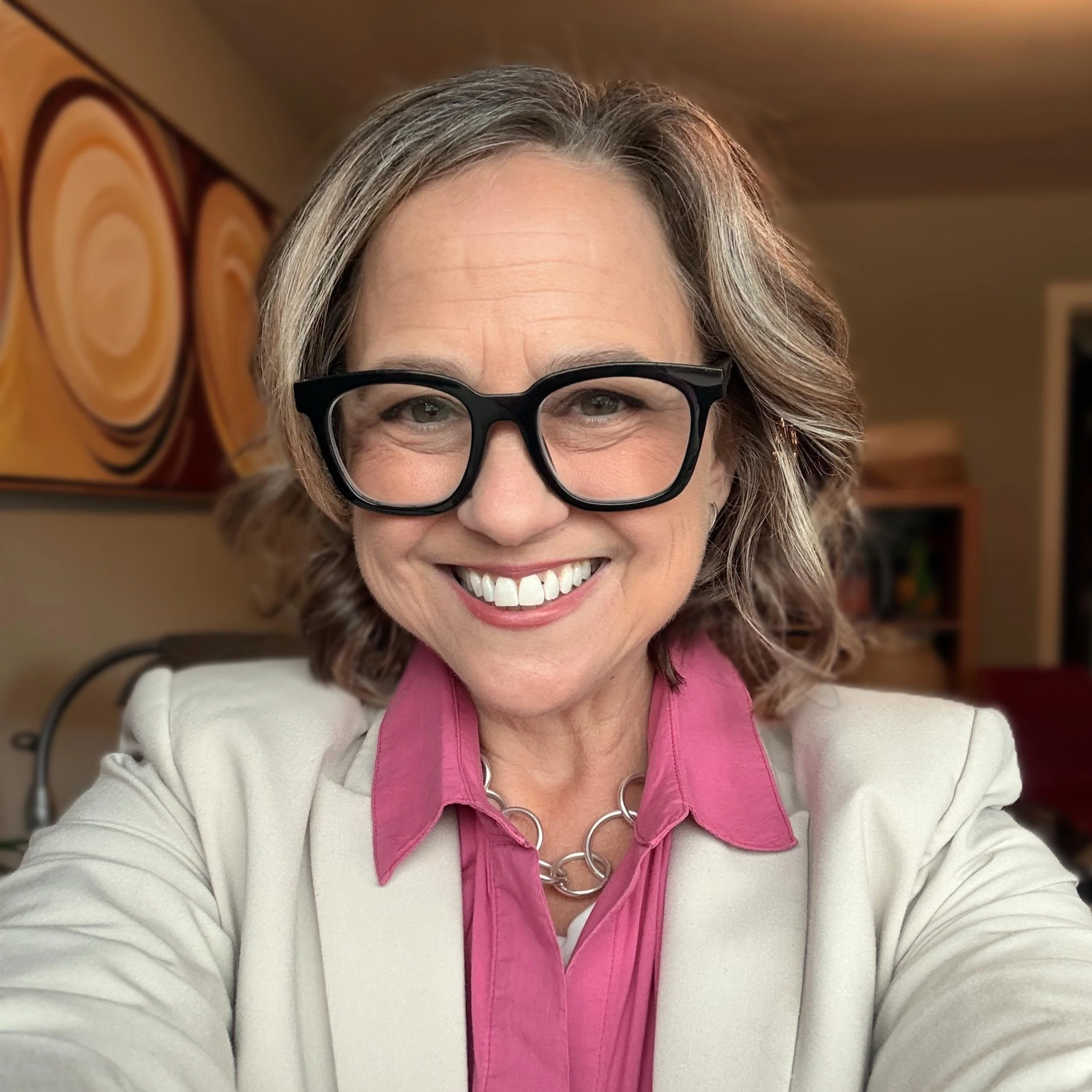 A middle-aged woman with shoulder-length blonde hair, wearing large black glasses, a white scarf, a black top, and a silver chain necklace, smiling outdoors against a dark blurred background.