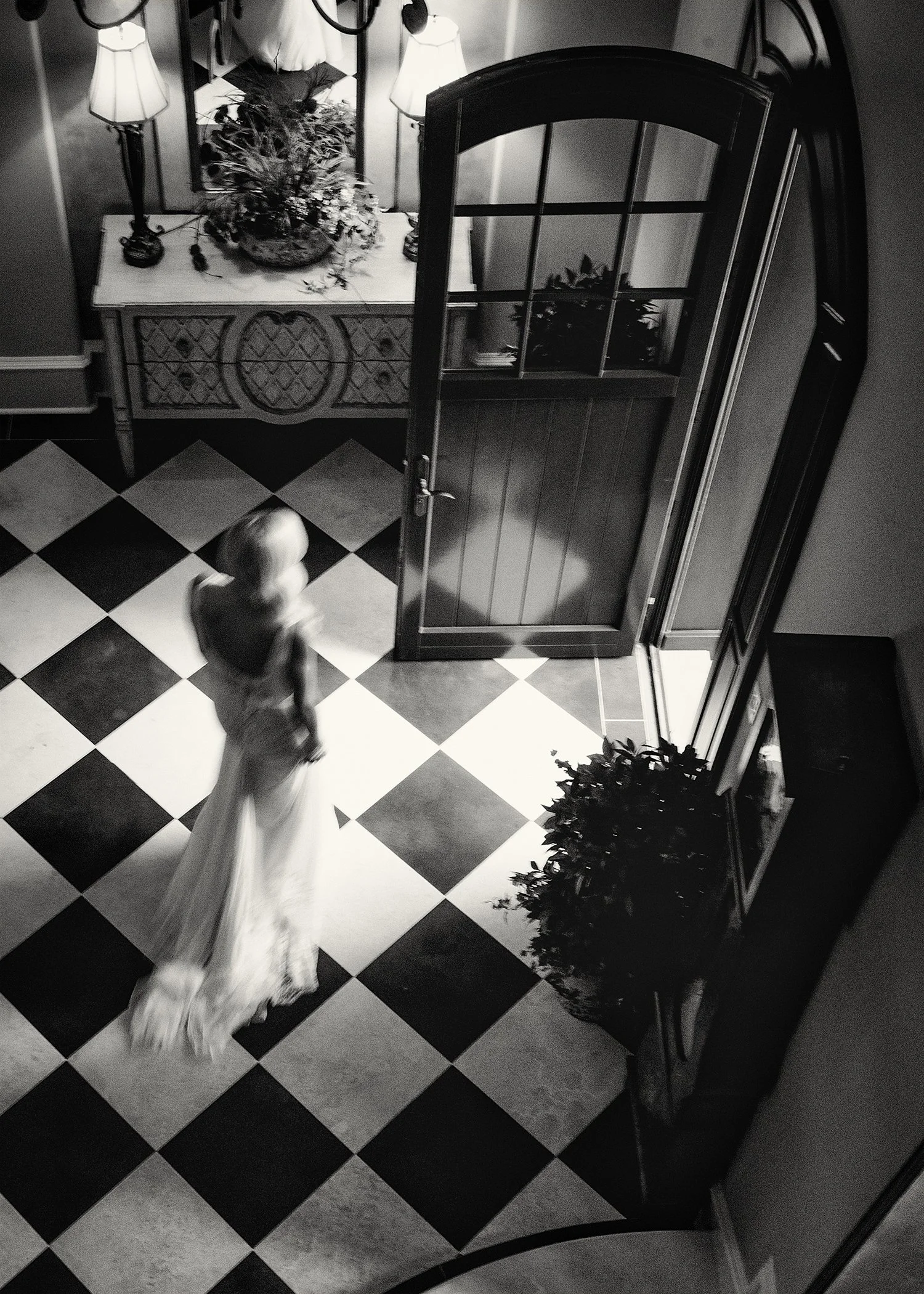 Black and white photo of an indoor scene with a checkered floor, a little girl in a long dress walking, a mirror on the wall, a small table with a flower arrangement, and a curved glass door