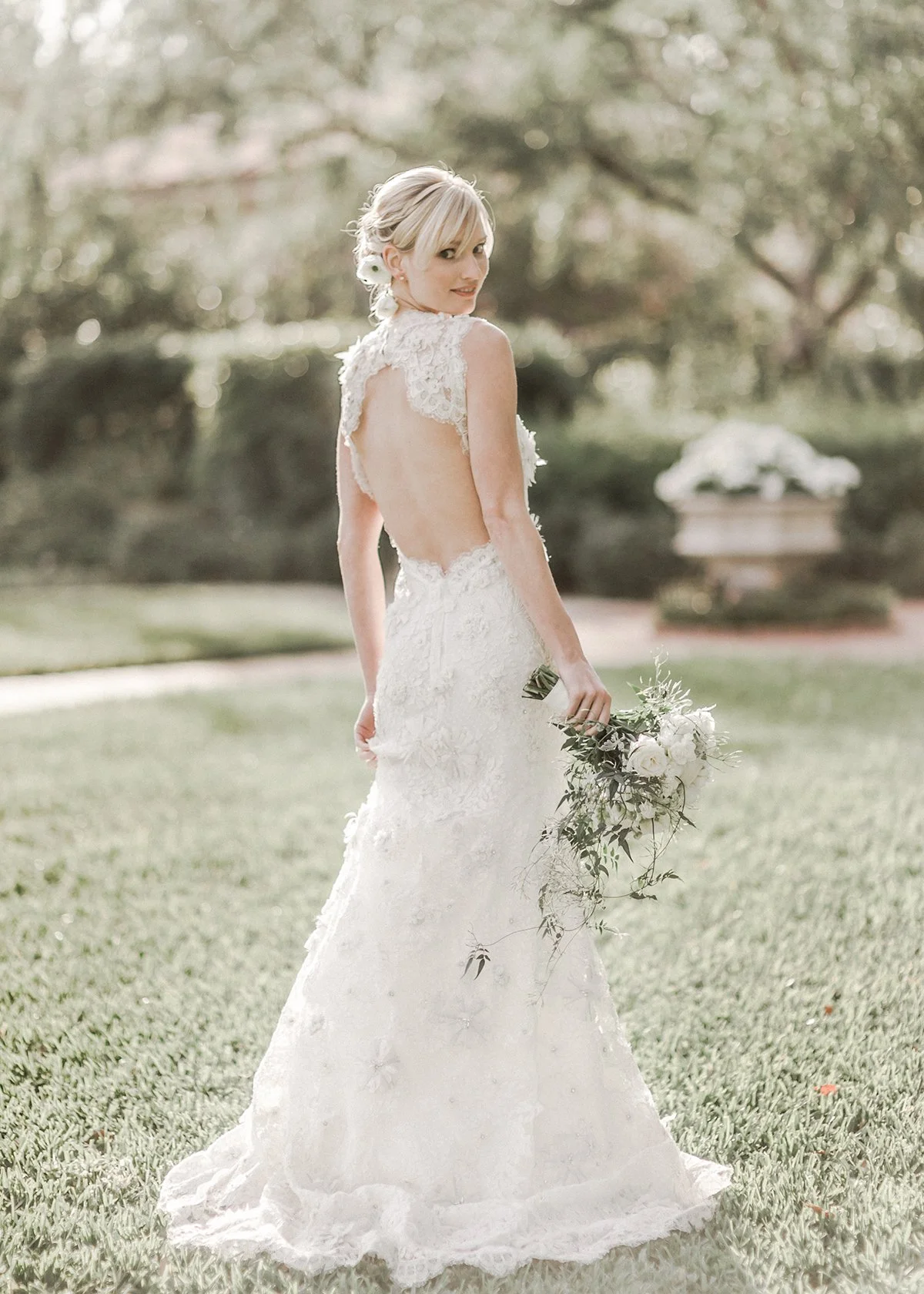 A bride in a white lace wedding gown standing on a lawn holding a bouquet of white flowers and greenery with her back slightly turned, outdoors with trees and a stone urn in the background.