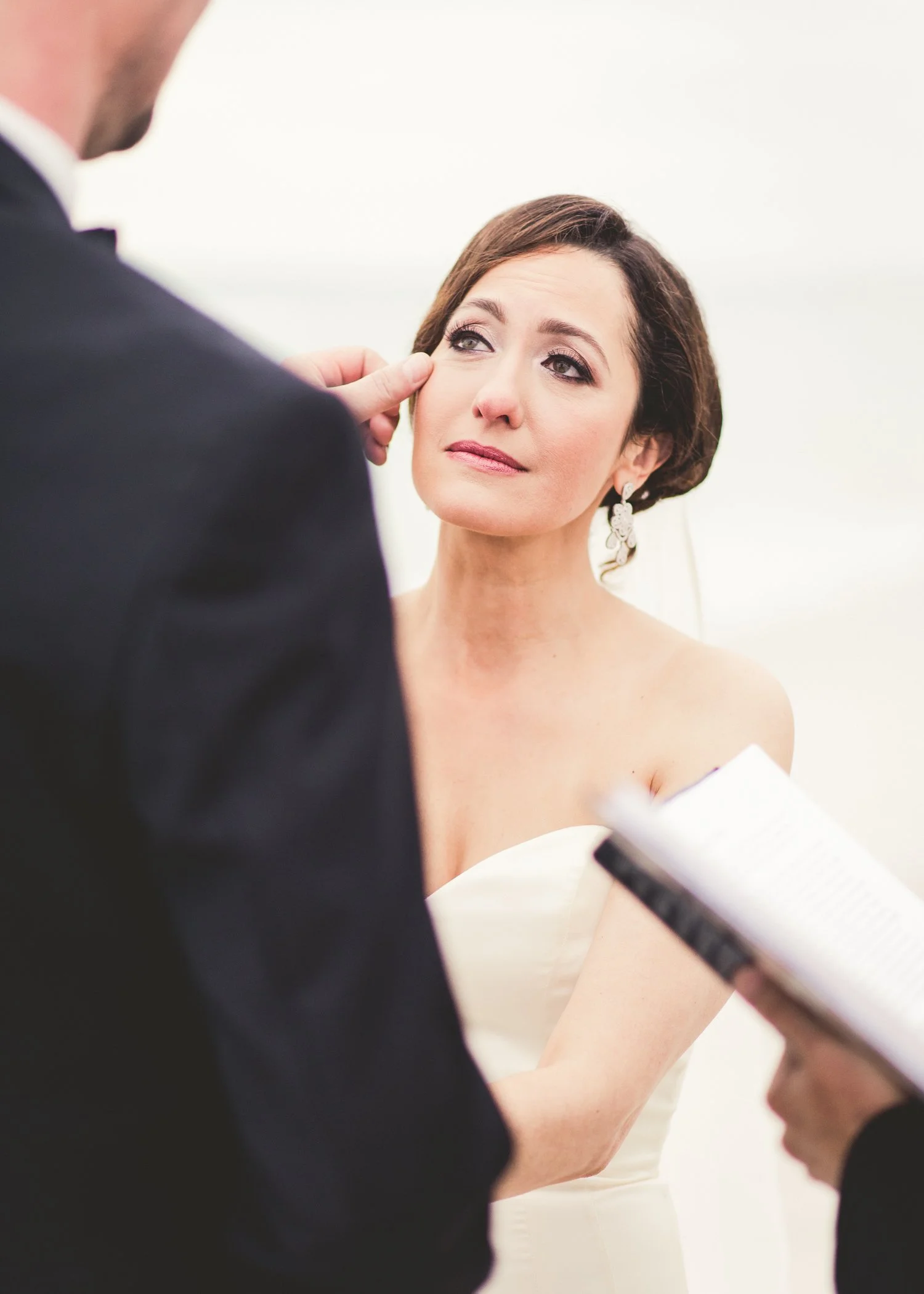 A bride with dark brown hair and earrings getting ready for her wedding, with an officiant or helper adjusting her dress or bouquet.