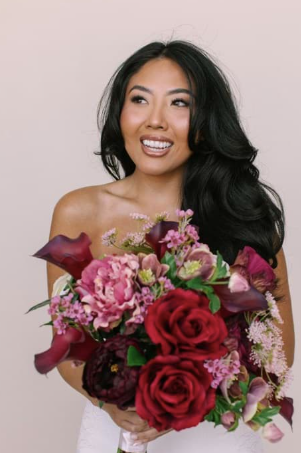 A woman with long black hair holding a large bouquet of pink, red, and purple flowers, smiling and looking to the side.