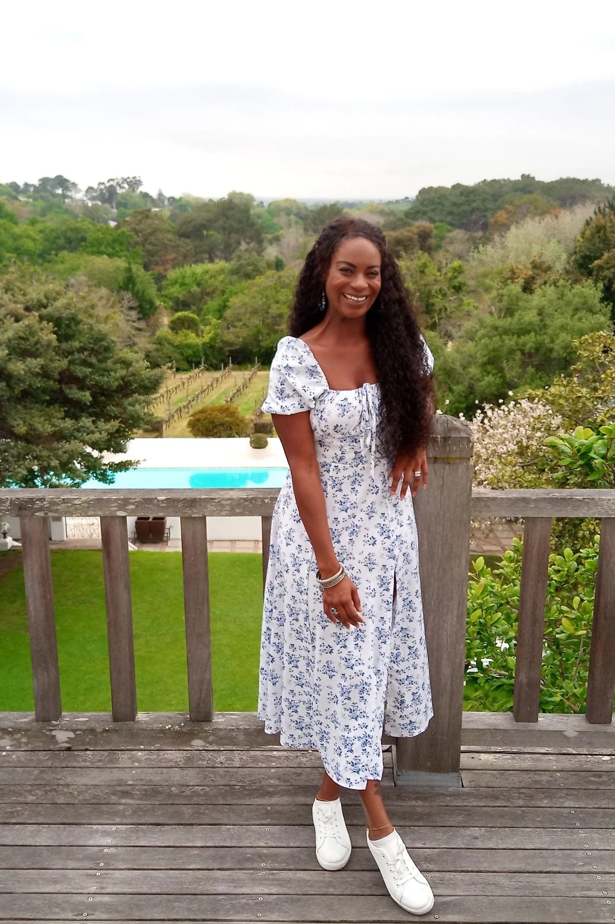 Smiling woman with long curly hair in a white dress with blue floral pattern standing on a wooden deck with a lush green landscape and pool in the background.