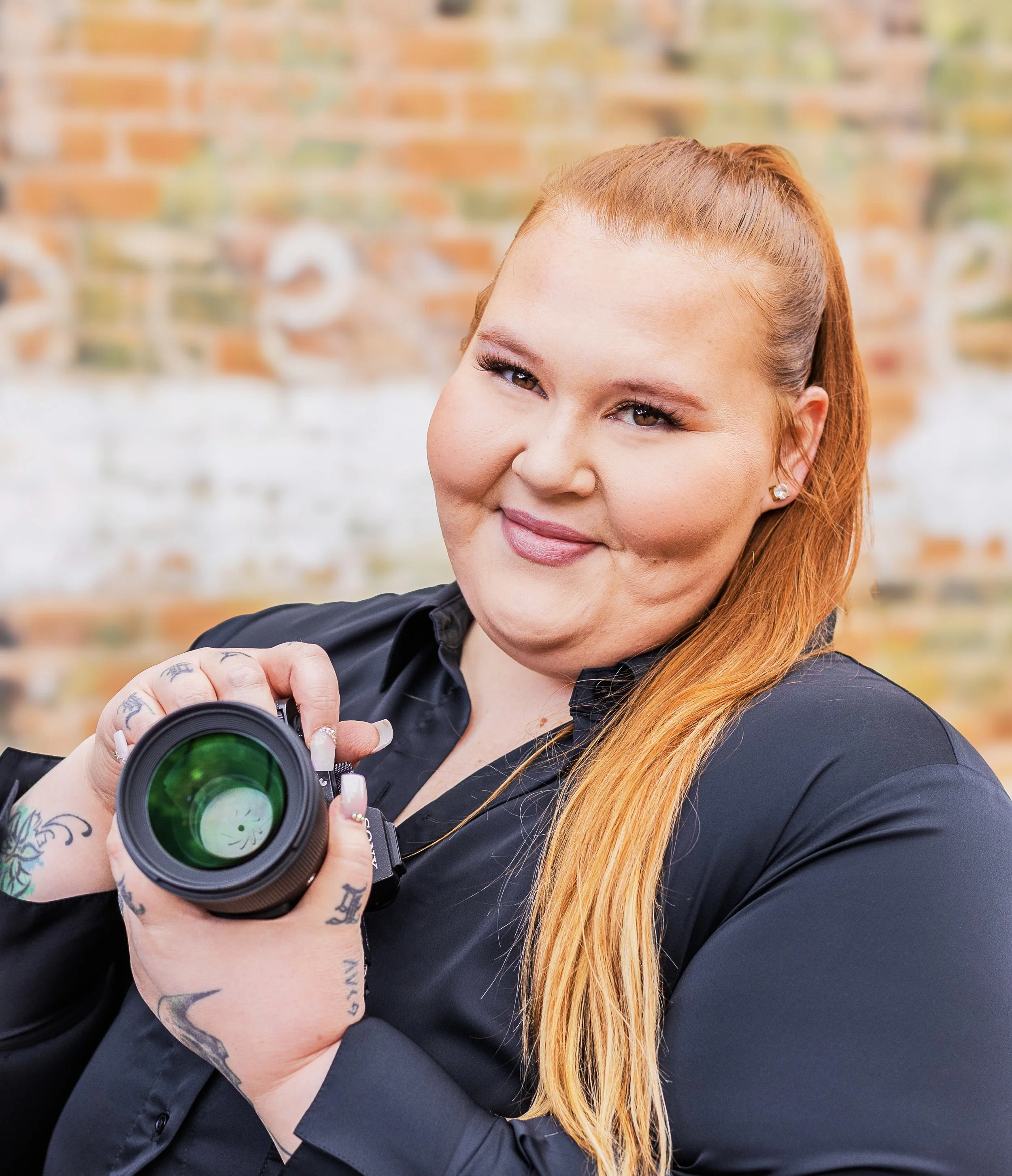A woman with long red hair and tattoos on her hand, holding a camera with a green lens, smiling at the camera with a brick wall background.