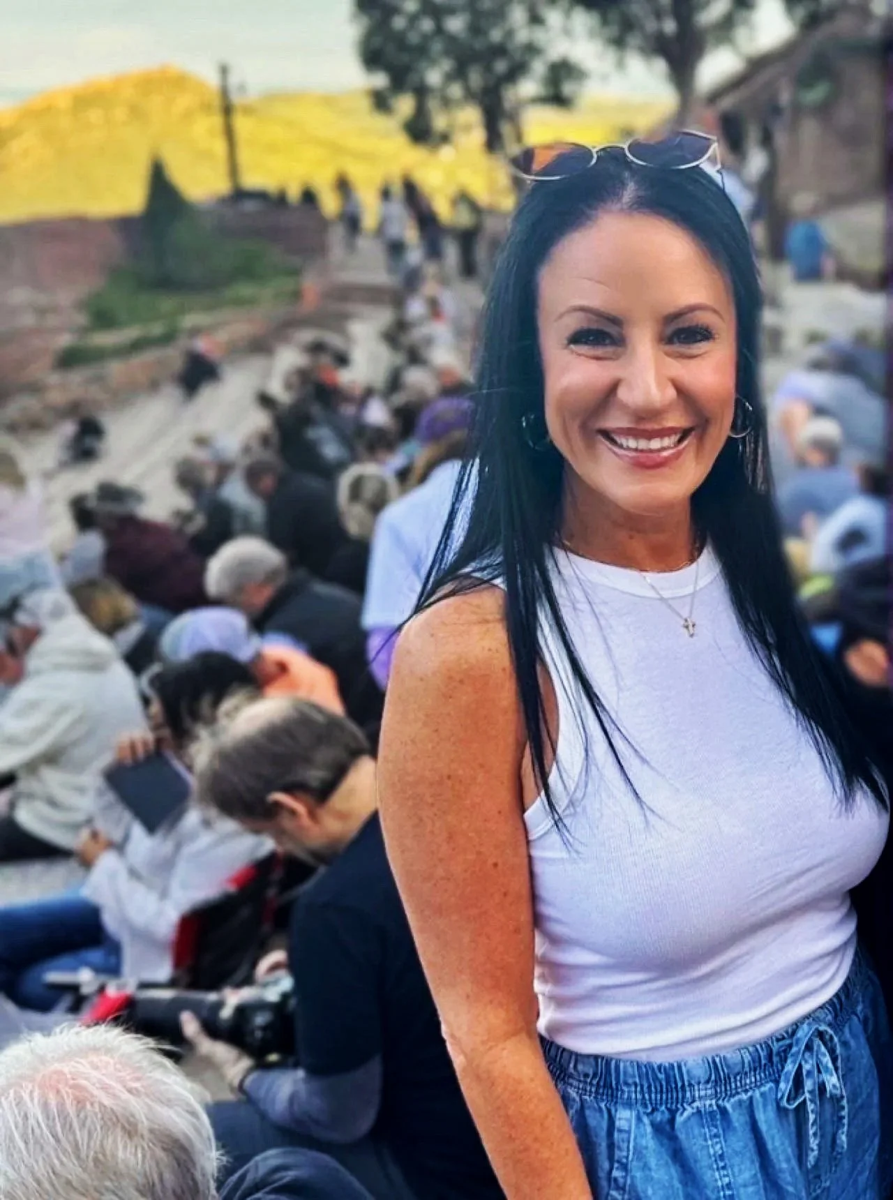 A woman with long black hair, wearing a white sleeveless top, denim shorts, sunglasses on her head, and a gold necklace, smiling at the camera outdoors with a large crowd sitting on steps behind her.
