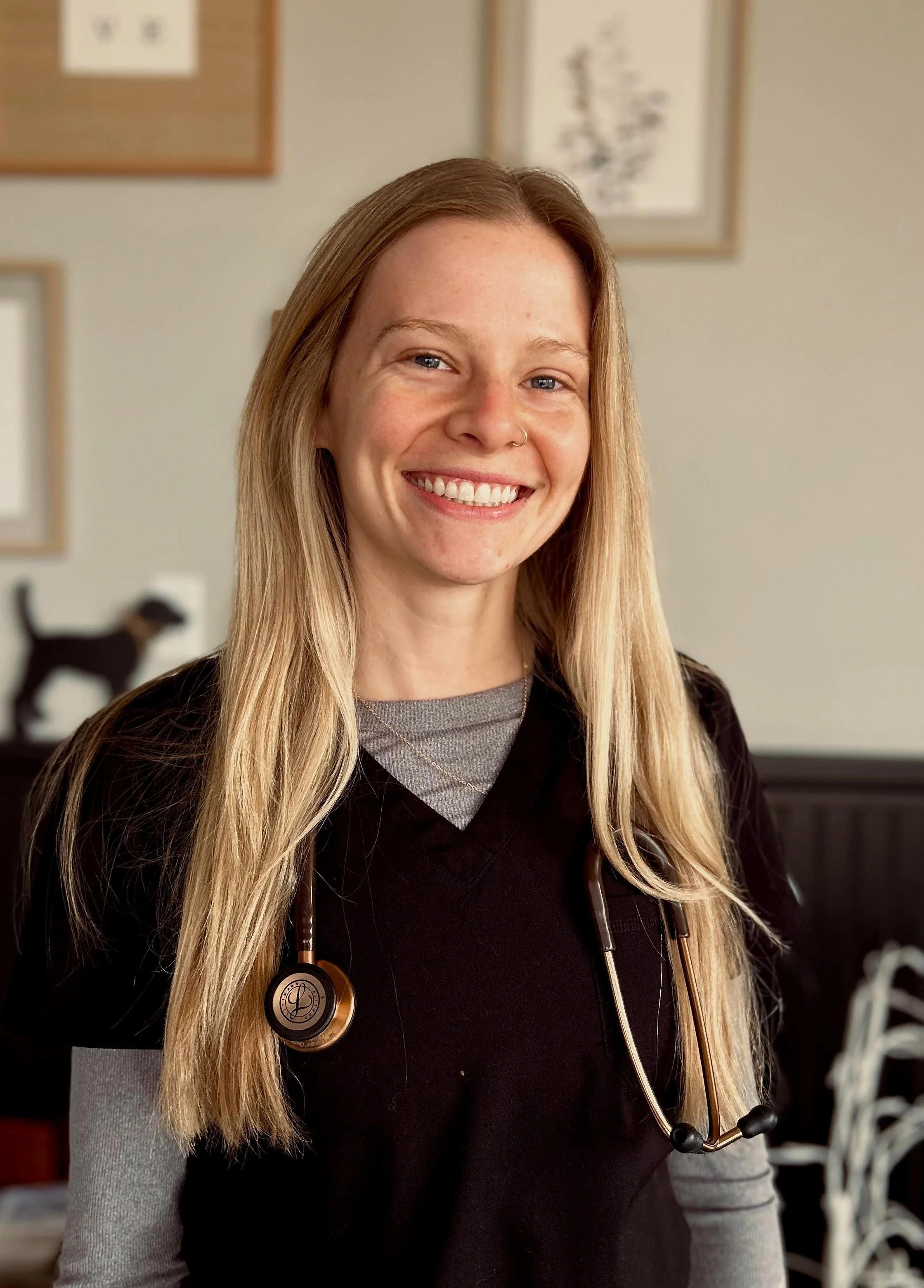 Smiling person wearing scrubs and a stethoscope, with framed pictures on the wall behind them.