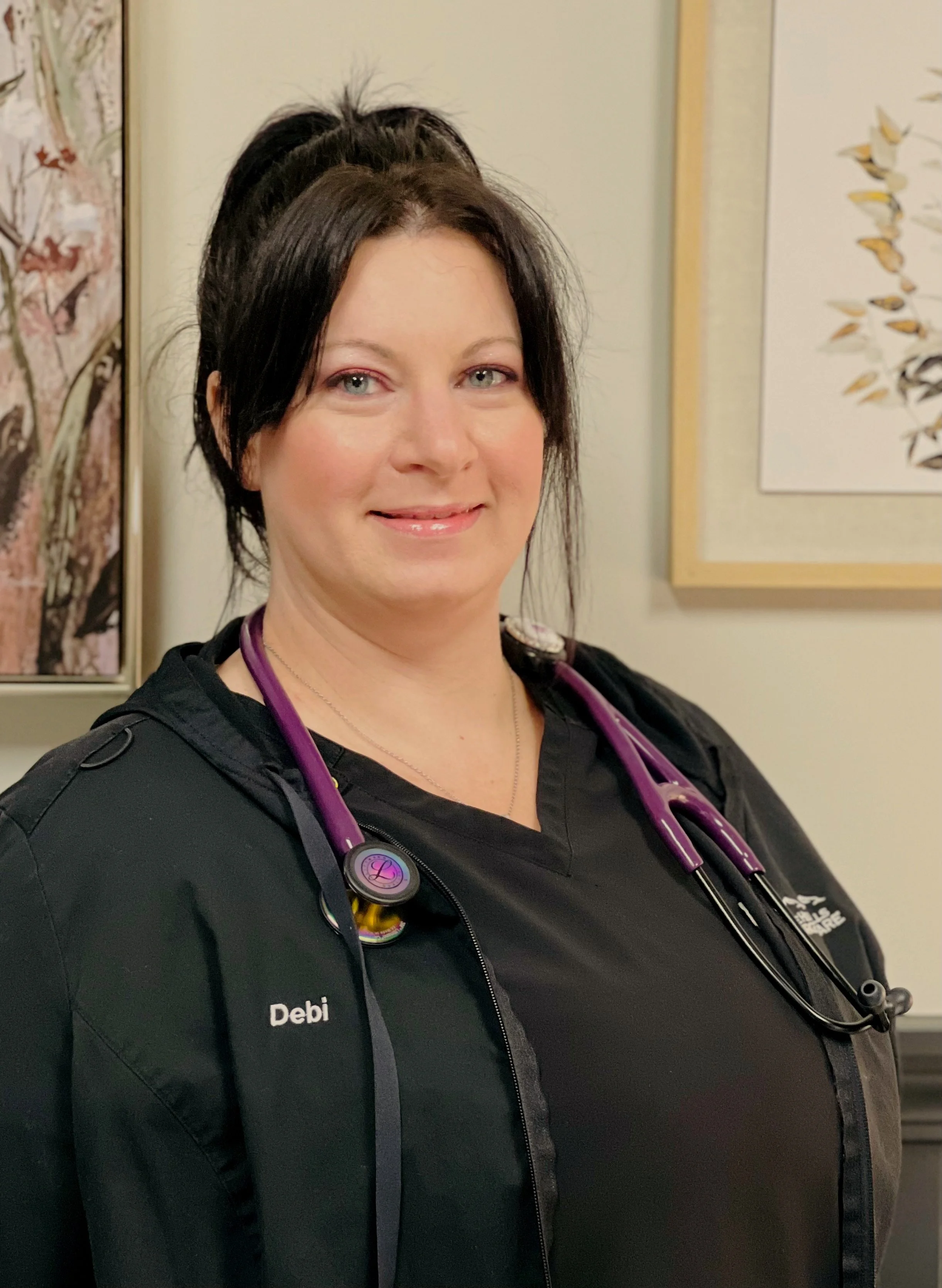 A woman in a black medical uniform with a stethoscope around her neck, standing indoors, smiling at the camera. Artwork is visible on the wall behind her.