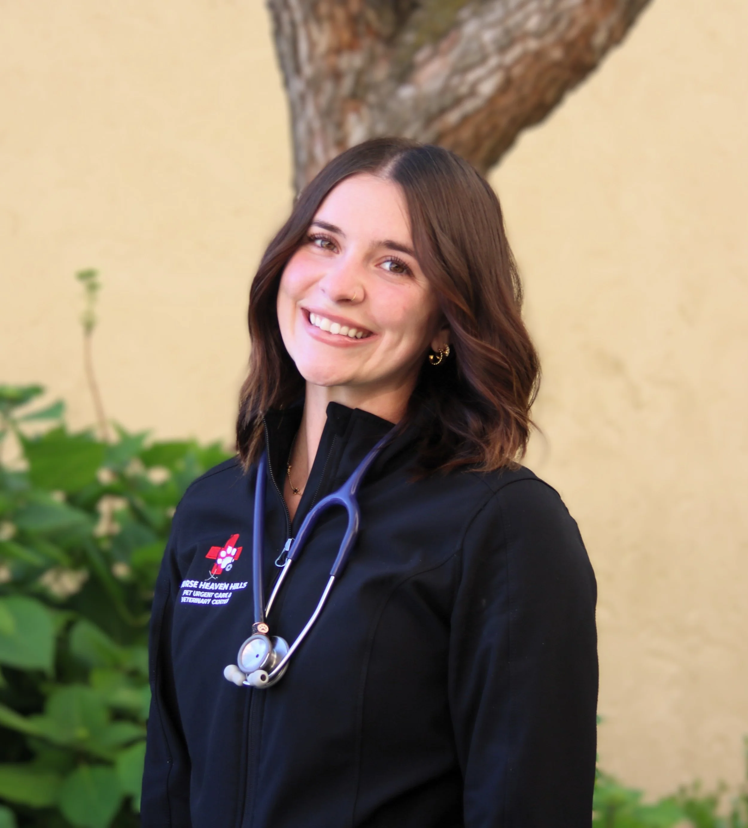 A smiling woman wearing a stethoscope and black scrubs with "Green Hills Pet Care" embroidered on them.