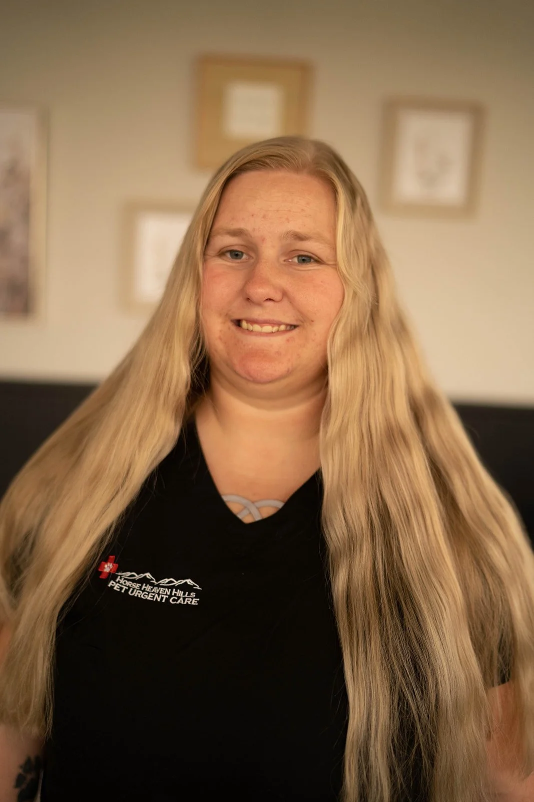 Person with long hair wearing a black "Horse Heaven Hills Pet Urgent Care" shirt, smiling in a room with framed pictures on the wall.