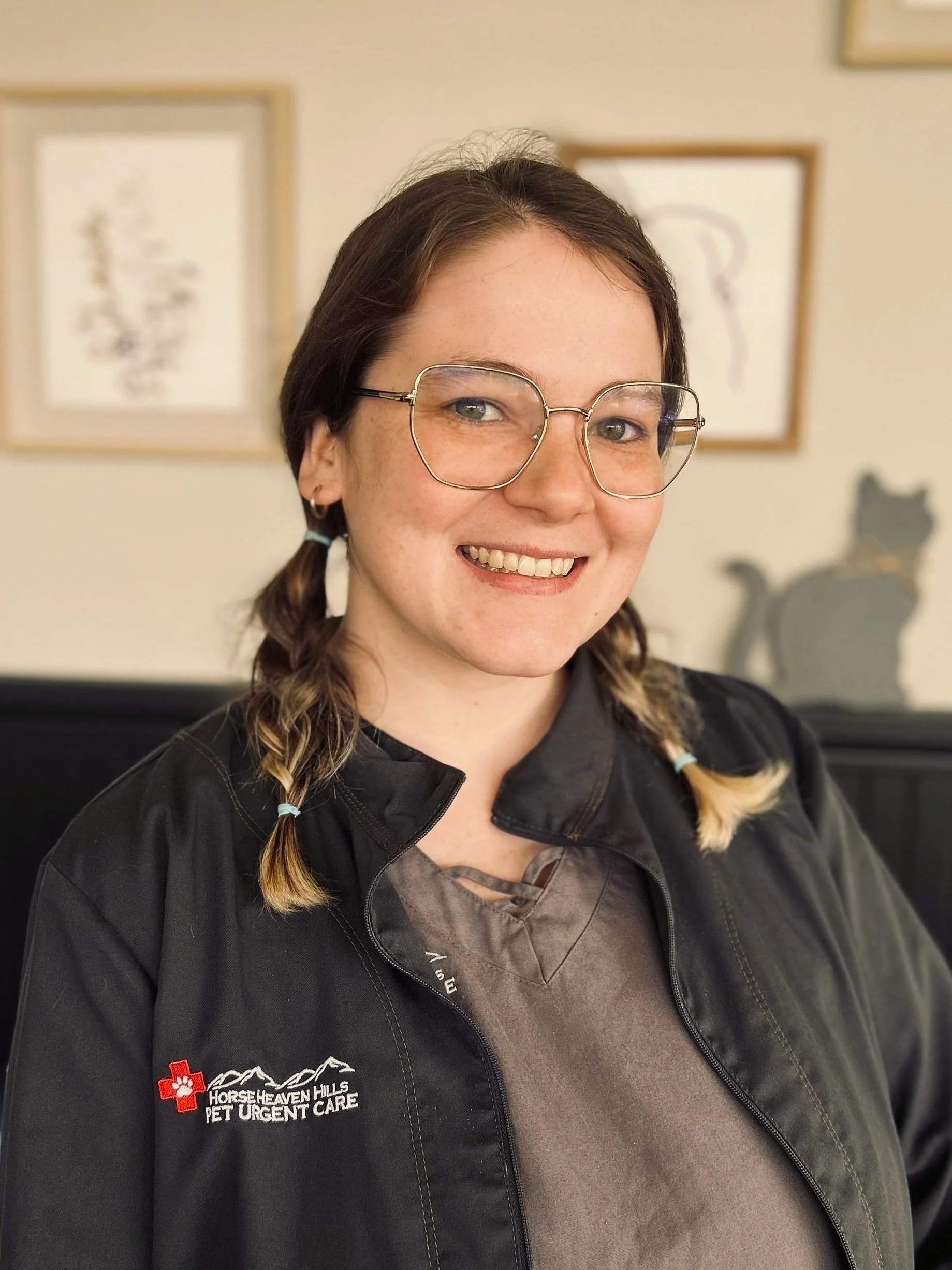 Person wearing glasses and a jacket with "Horse Heaven Hills Pet Urgent Care" logo, smiling indoors, with framed art and cat silhouette in the background.