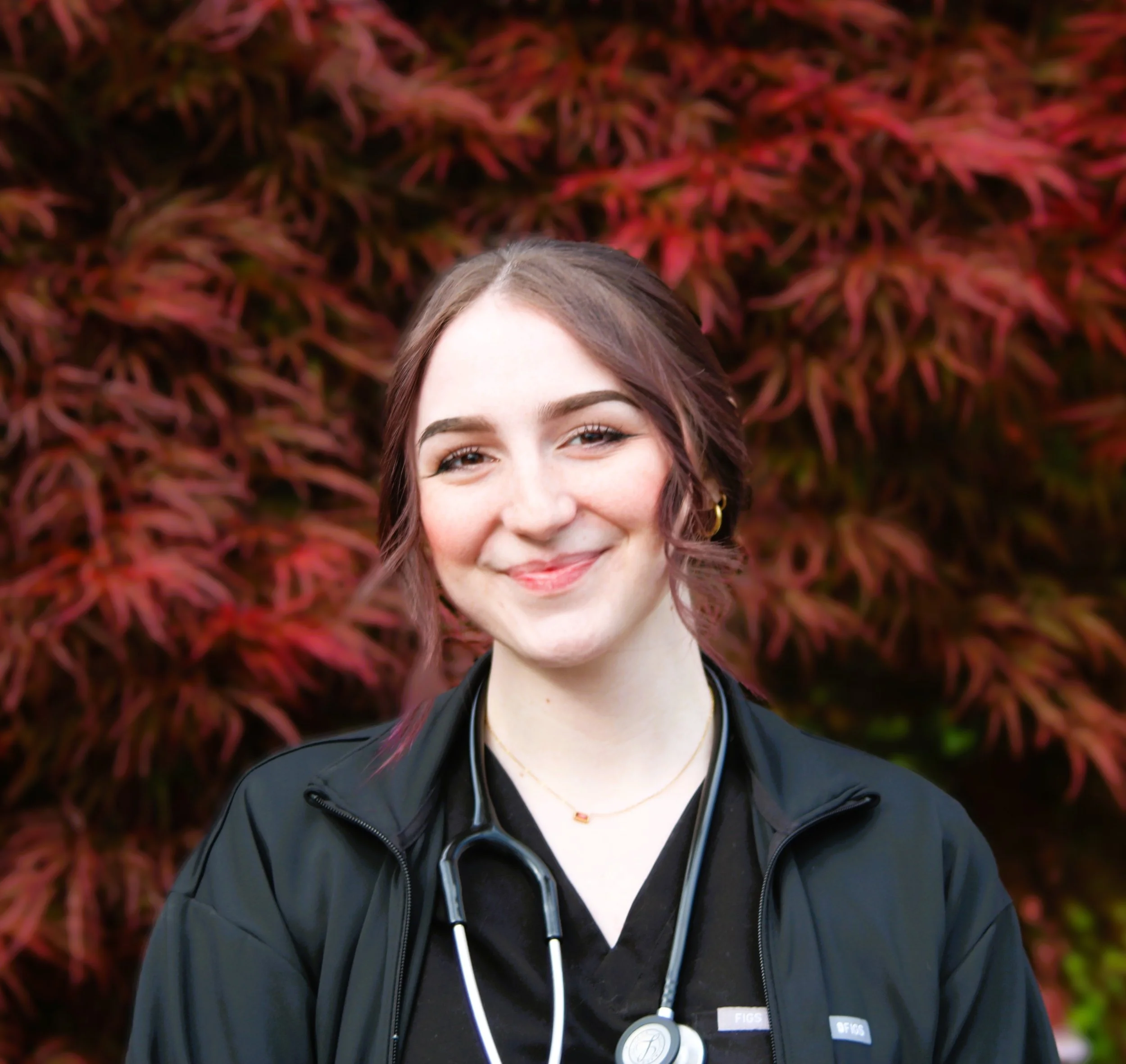 Woman wearing a black scrubs top with a stethoscope, standing indoors.