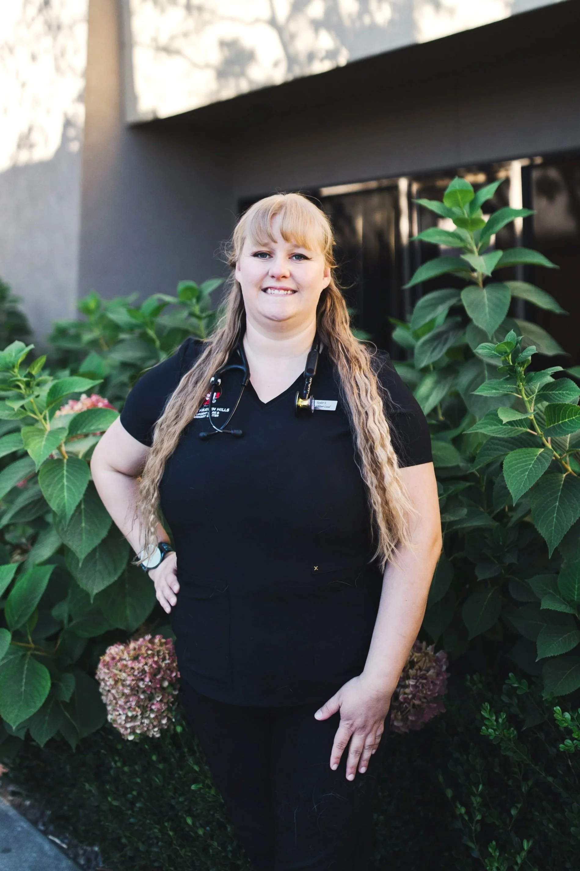 Person with long hair wearing a black "Horse Heaven Hills Pet Urgent Care" shirt, smiling in a room with framed pictures on the wall.