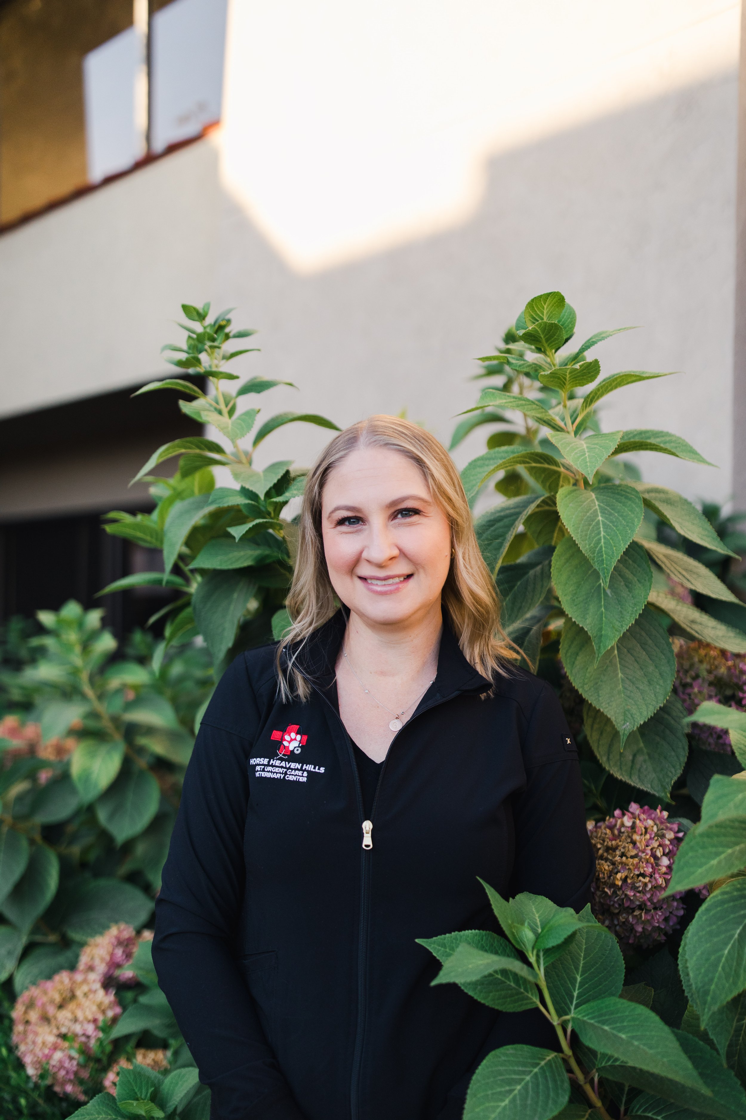 Person with glasses in a forest, wearing black long-sleeve shirt and jewelry, standing with arms crossed.